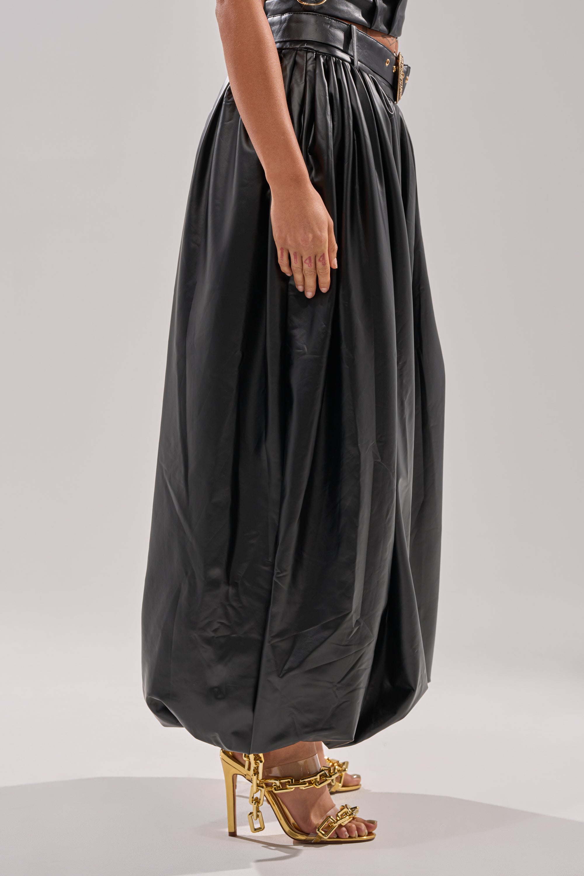 Wearing the ZONA BALLOON LEATHER TROUSER, a person stands against a plain light background paired with gold high-heeled sandals featuring chain details.