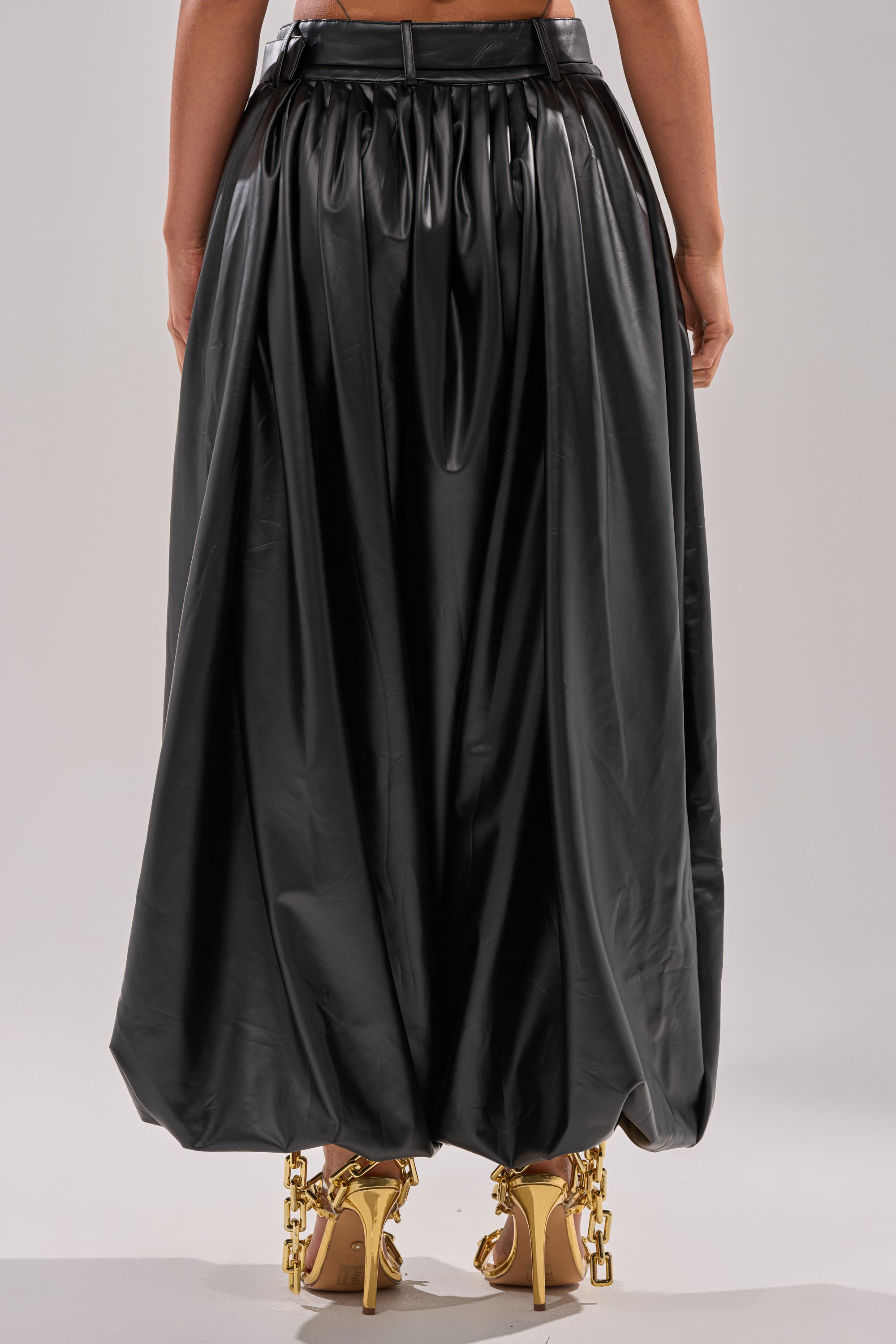 A person stands facing away, wearing the ZONA BALLOON LEATHER TROUSER—a shiny black balloon-style piece—paired with gold high-heeled sandals adorned with chunky chain details.