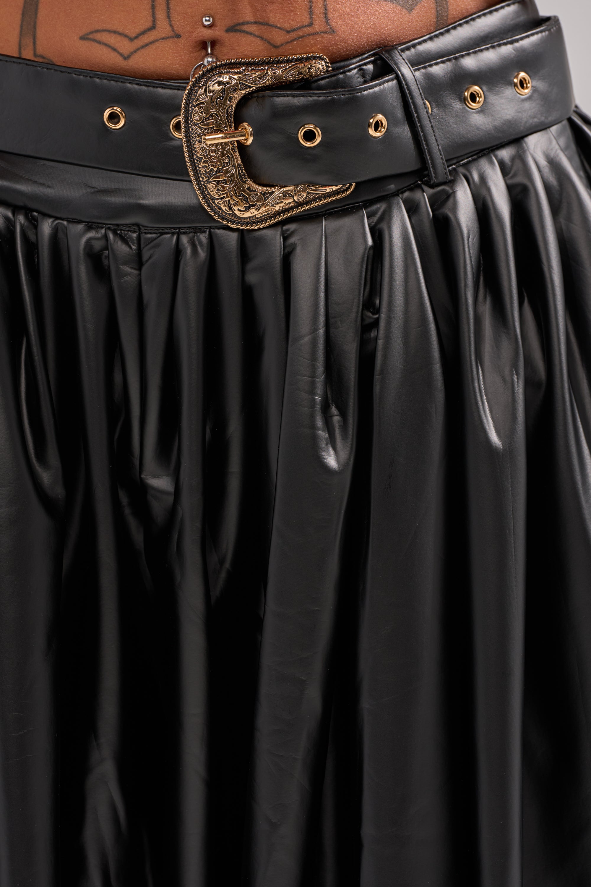 A person wears the ZONA BALLOON LEATHER TROUSER with a black belt featuring a large gold buckle, echoing the sleek look of pleated skirts. A tattoo peeks out above the belt on their lower abdomen.