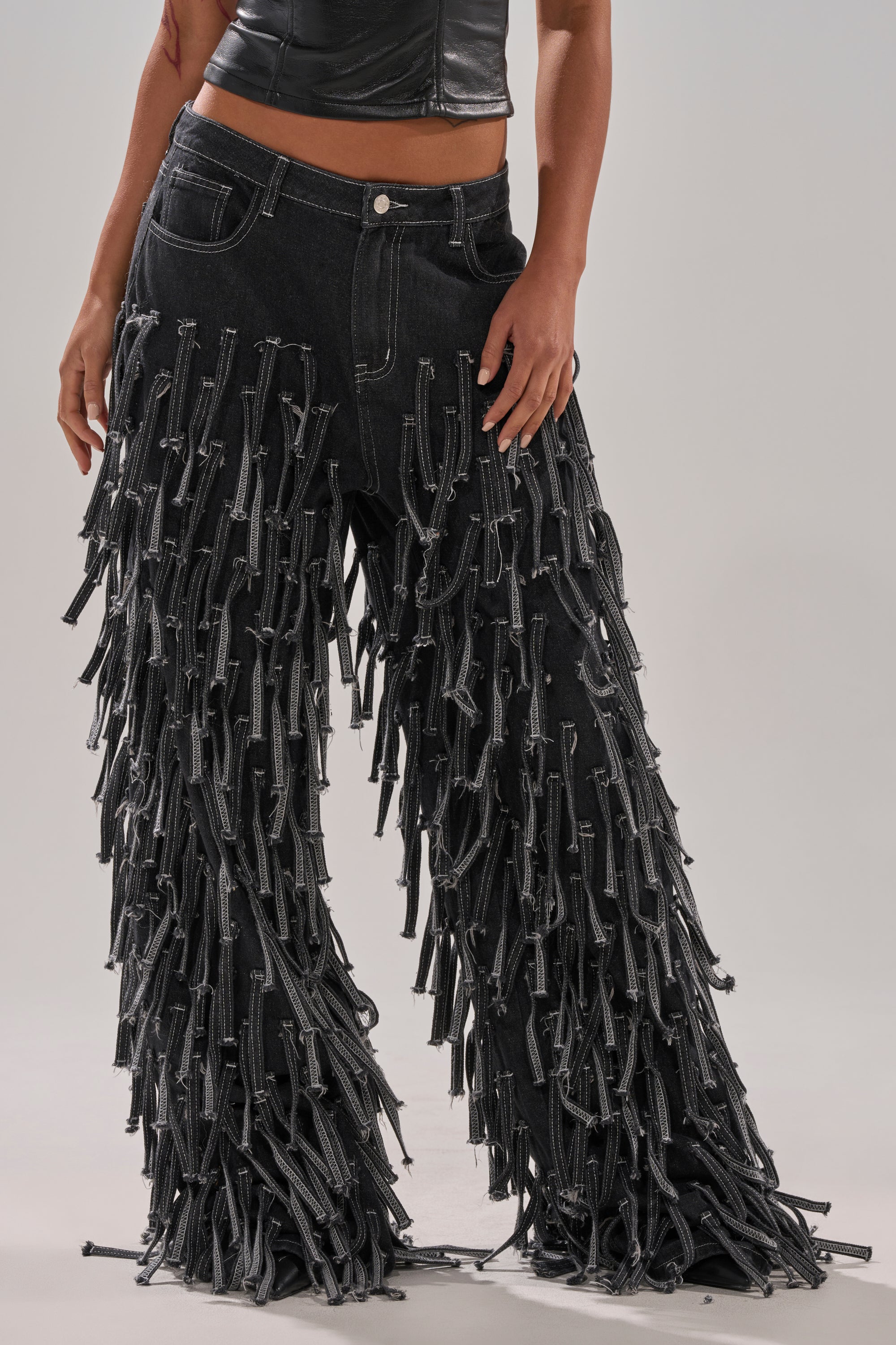 A person wears CRAZY FOR YOU FRINGE DENIM, high-rise black pants with fringe and zipper tassels, paired with a cropped black leather top; the image is cropped above the shoulders.