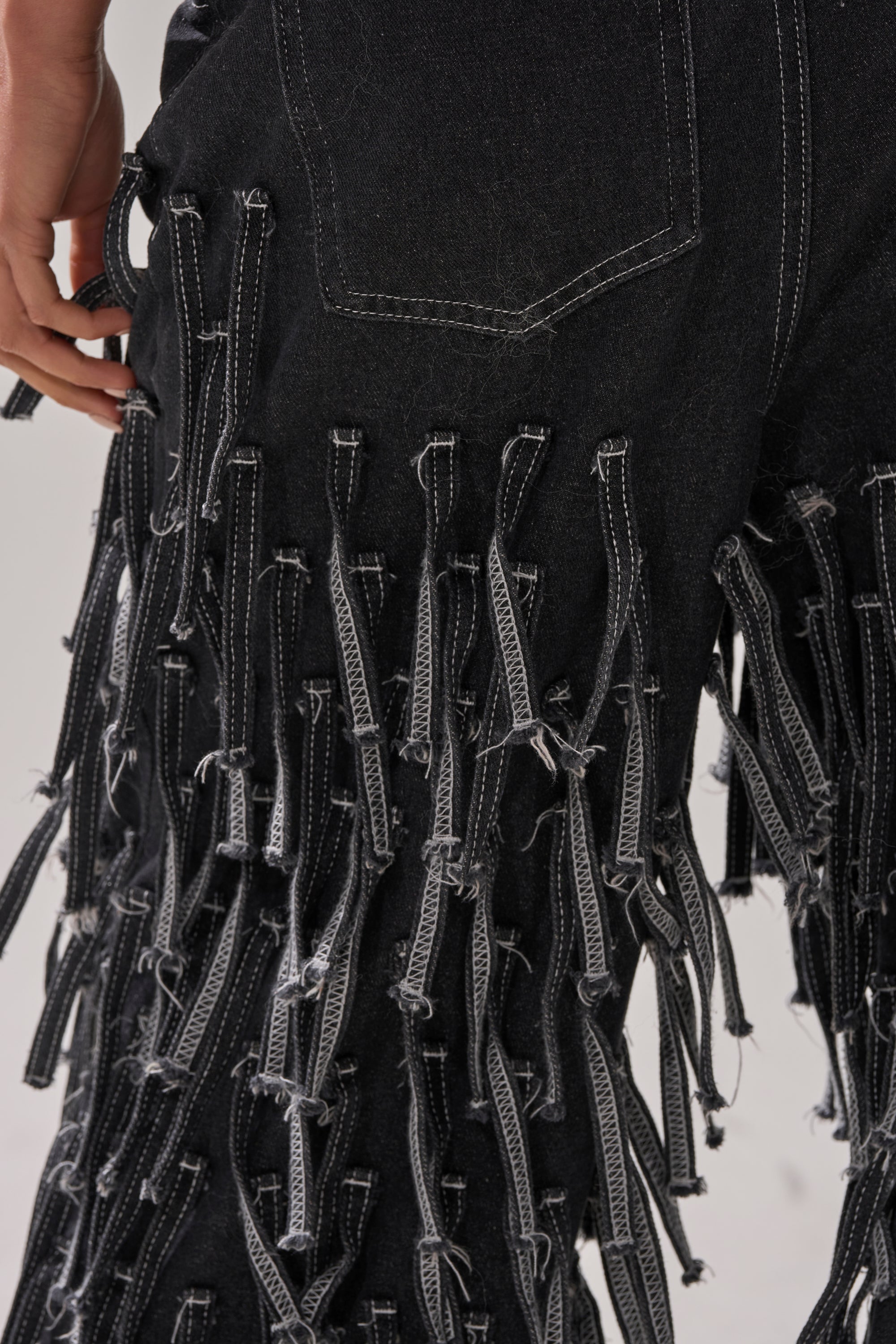 Close-up of CRAZY FOR YOU FRINGE DENIM shows black wide-leg jeans with white stitching and rows of loose fringe strips for a textured look. A hand is visible near the upper left corner.