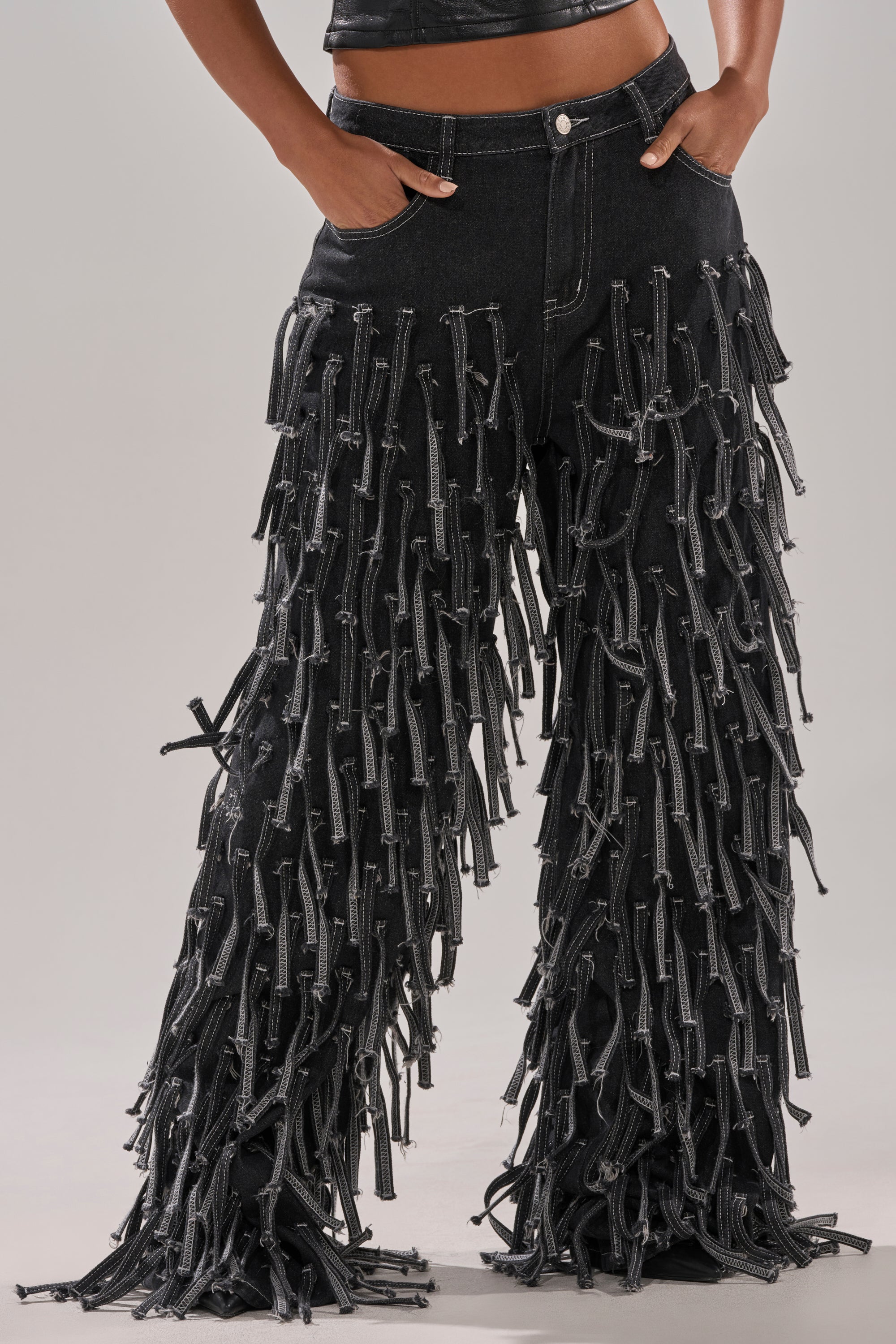 A person wearing the CRAZY FOR YOU FRINGE DENIM: black high-rise, wide-leg jeans with dramatic hanging fringe strips and a zipper fly, standing with hands in pockets.