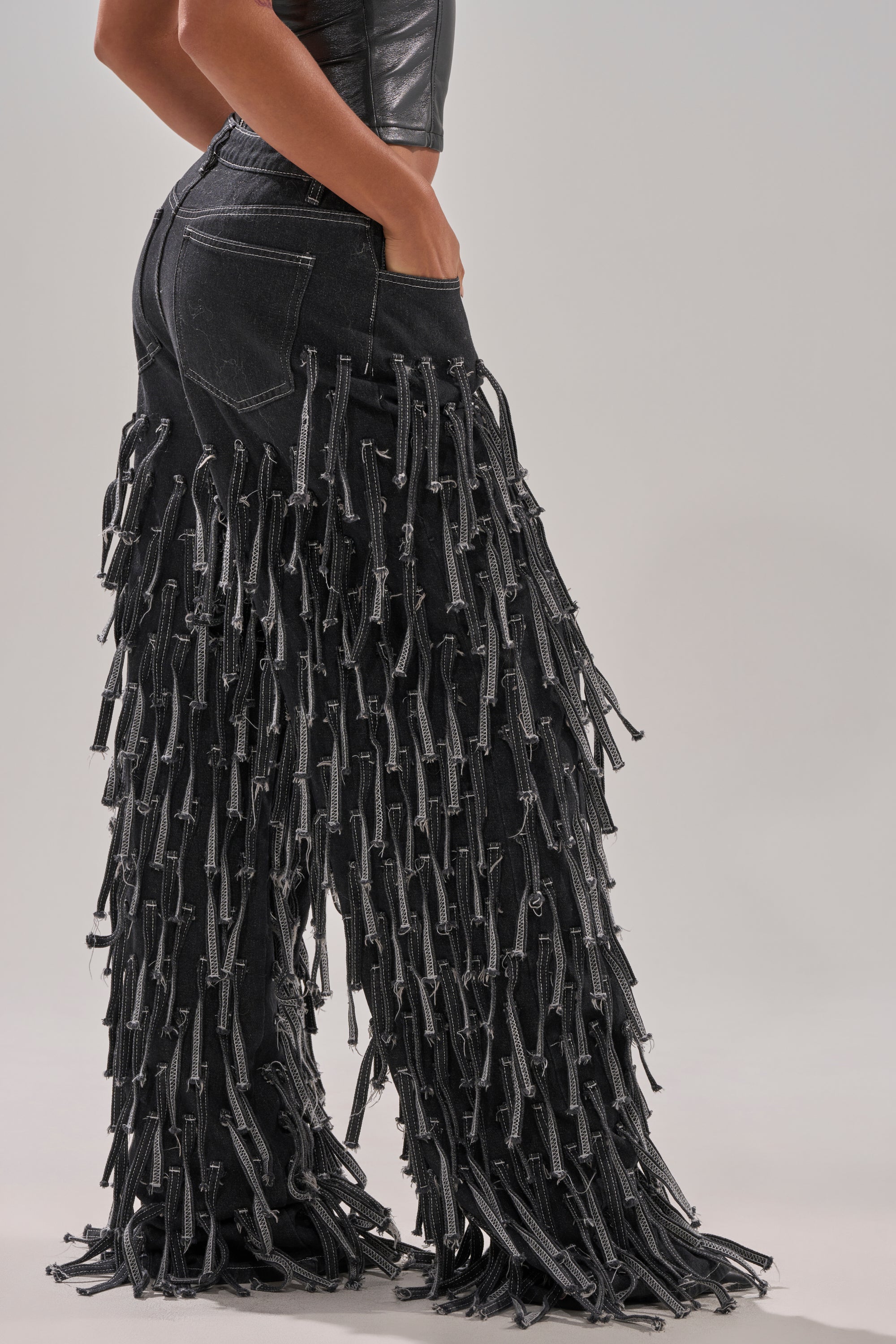 Wearing the CRAZY FOR YOU FRINGE DENIM, a pair of black high-rise wide-leg jeans with long, frayed tassels, this person stands hands-in-pockets and pairs them seamlessly with a black top.