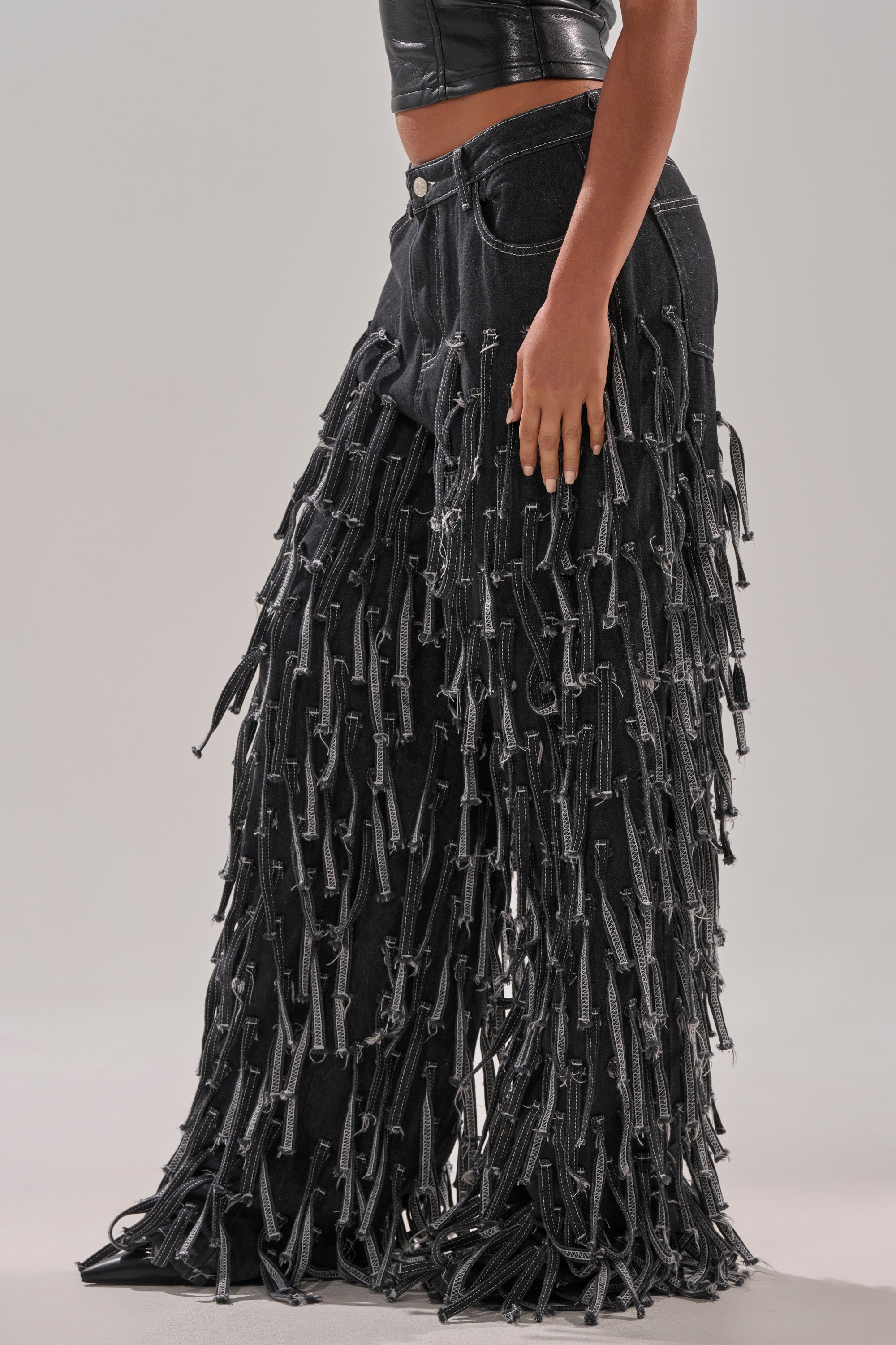 Wearing the CRAZY FOR YOU FRINGE DENIM black wide-leg jeans with zipper tassels and a black cropped leather top, this person stands against a plain background for a bold fringe denim look.