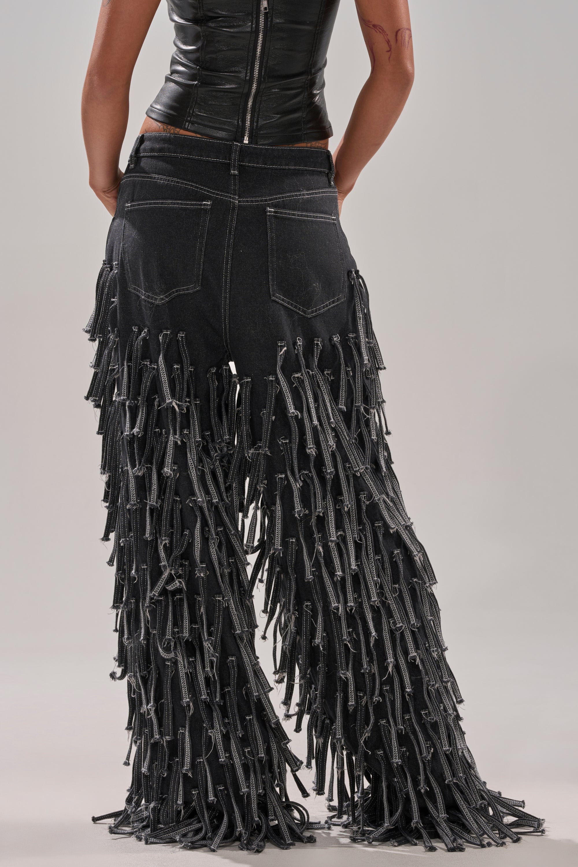 A person faces away, wearing the CRAZY FOR YOU FRINGE DENIM—high-rise black jeans with long denim fringe—and a sleeveless black zip-up top. The background is plain and light-colored.