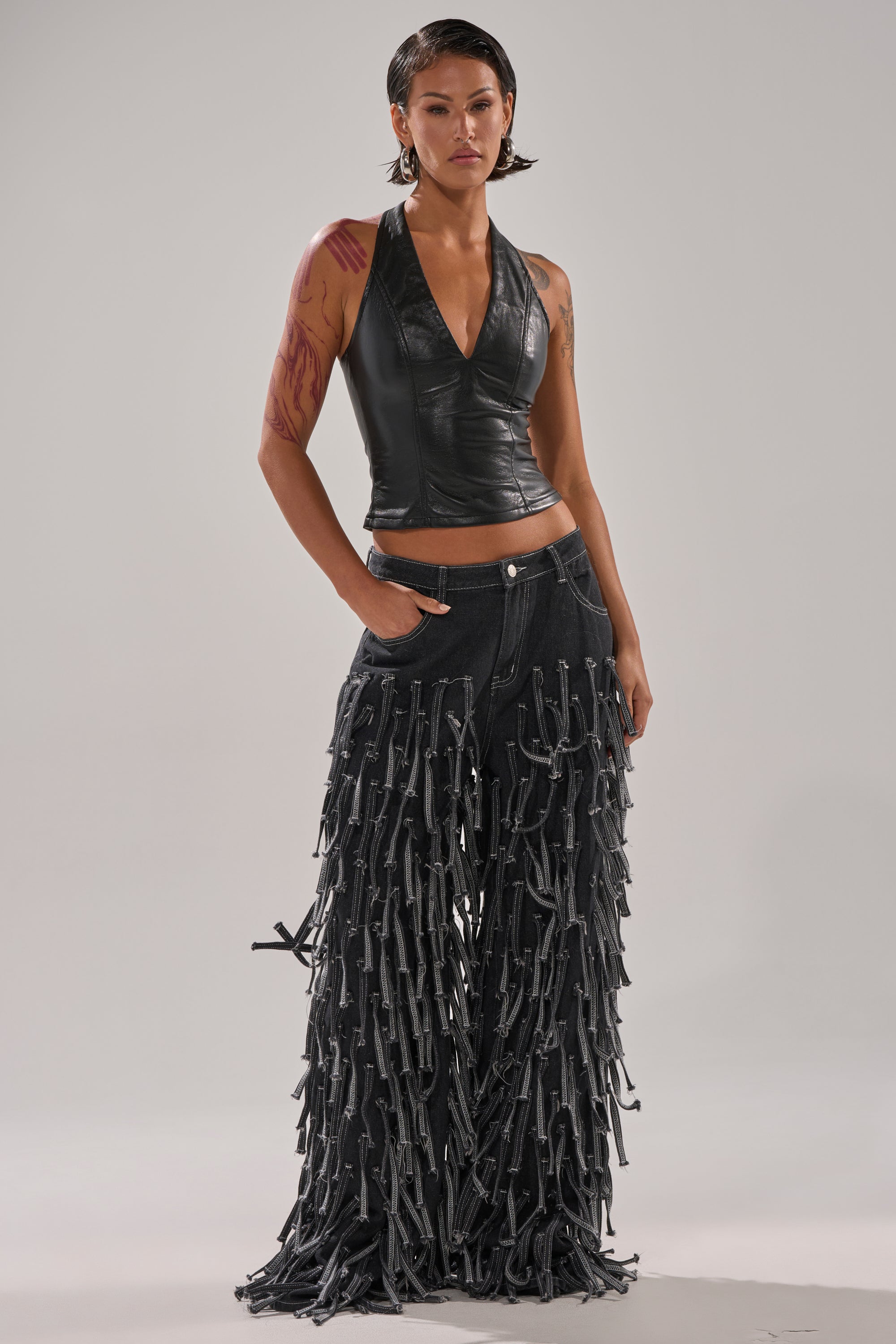 A person with short, slicked-back hair wears a black leather halter and the CRAZY FOR YOU FRINGE DENIM jeans, standing with one hand in a pocket against a simple light background.