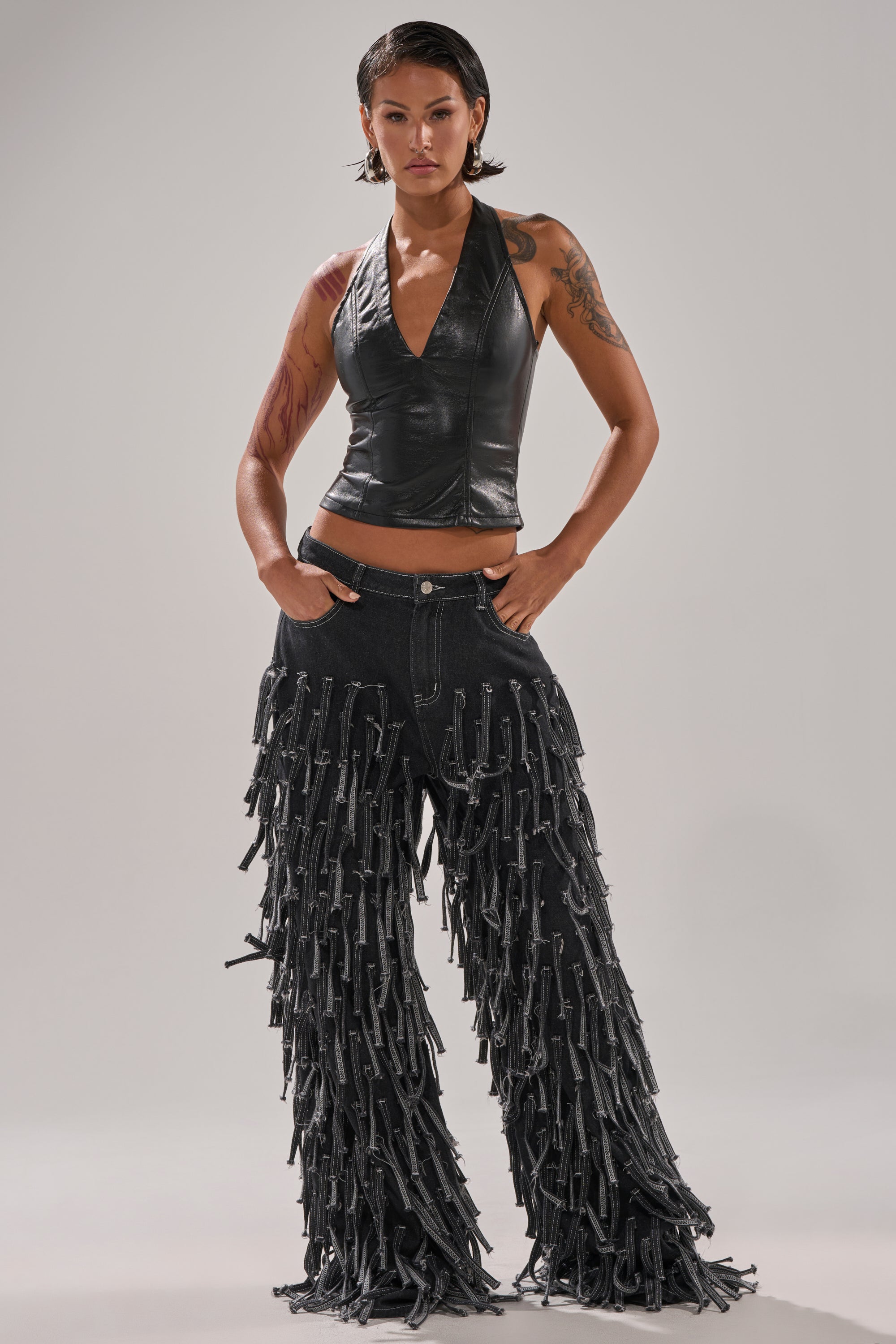 A person stands confidently in the CRAZY FOR YOU FRINGE DENIM jeans and a black faux leather halter top, displaying arm tattoos against a plain background.