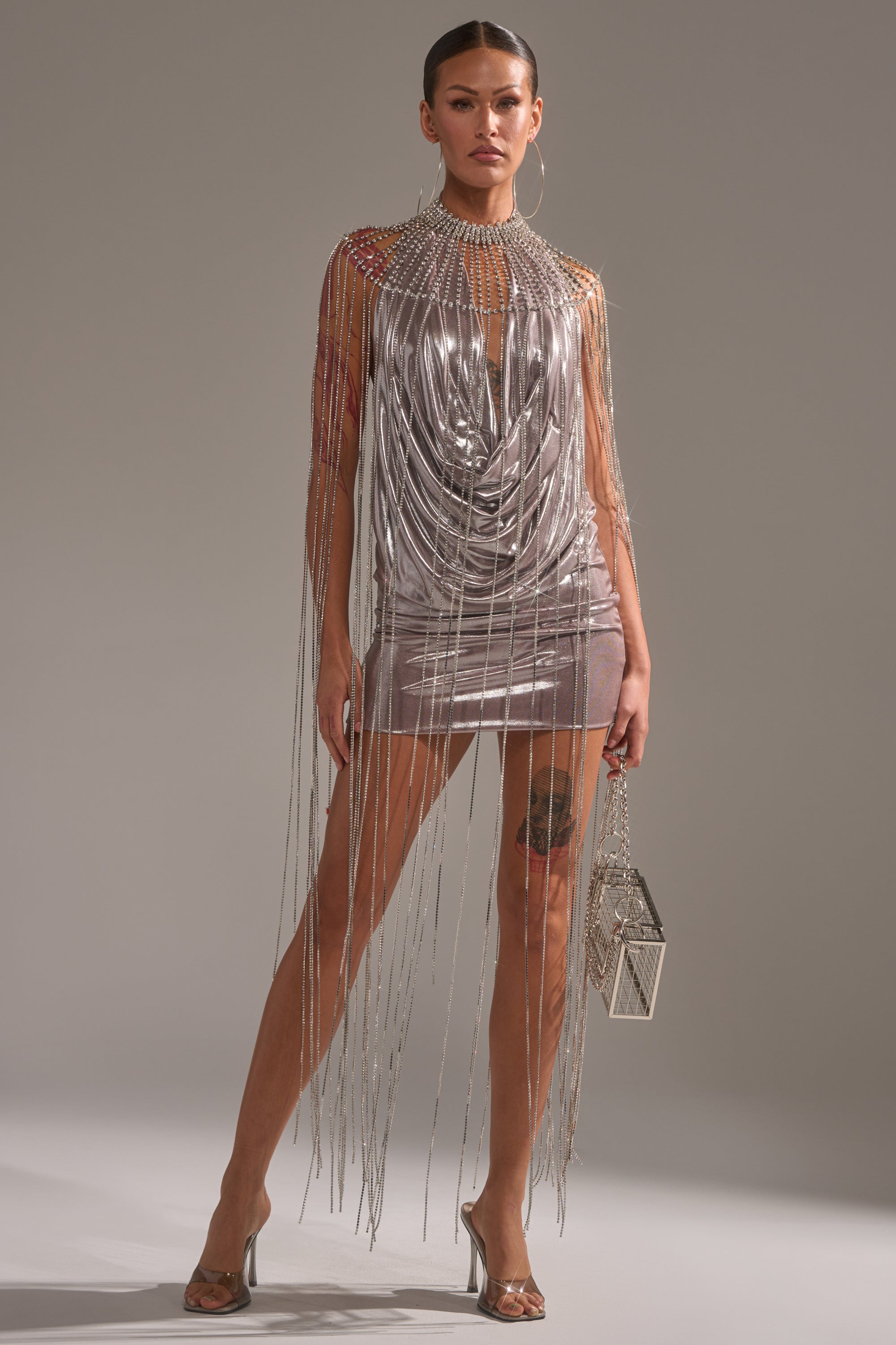 A woman stands confidently in a metallic silver mini dress with rhinestone fringe, clear heels, a geometric purse, hoop earrings, and her hair pulled back, wearing the TRU STATEMENT NECKLACE as her standout accessory. 7