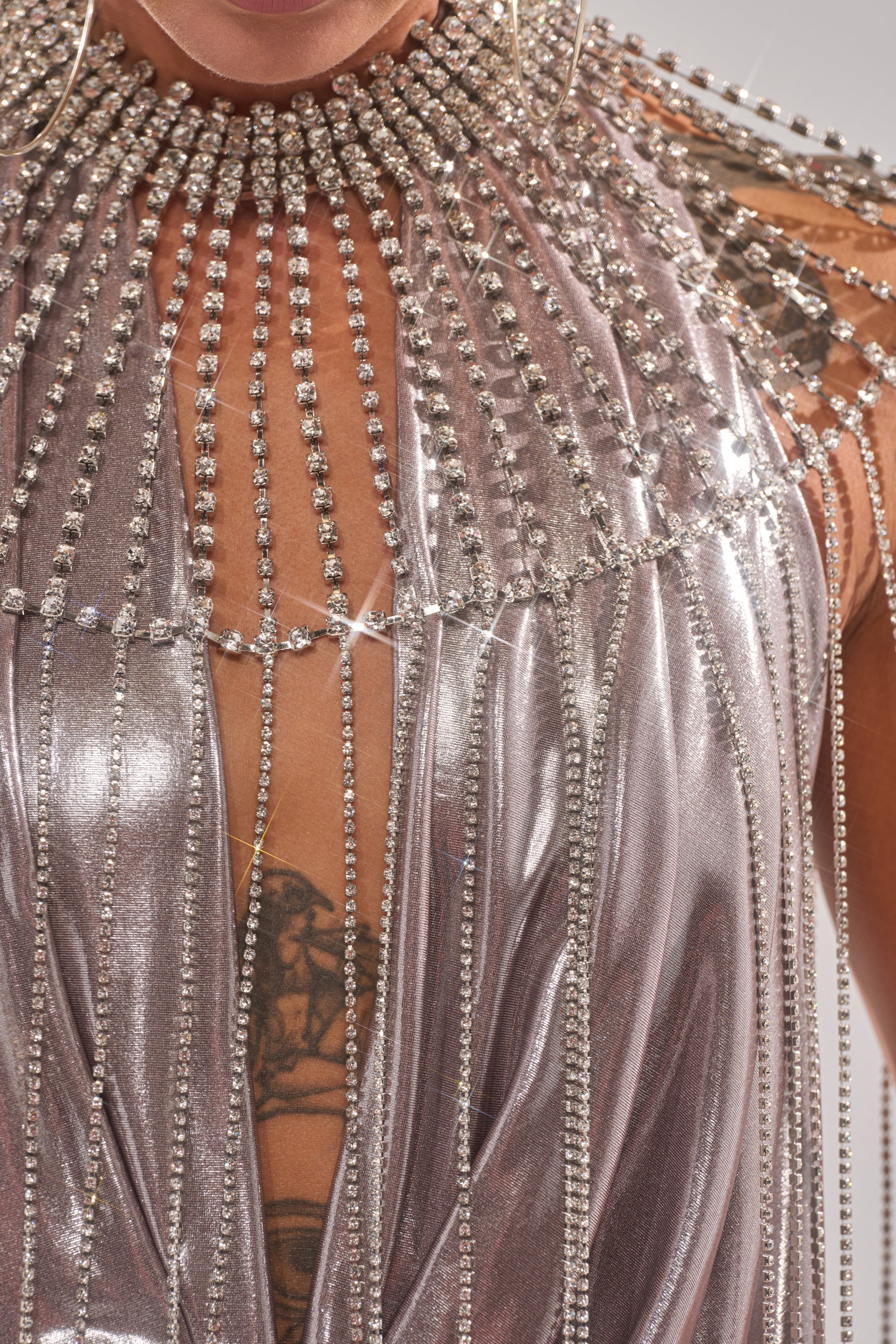 A close-up of a person wearing a metallic silver outfit with rhinestones and beads, paired with the TRU STATEMENT NECKLACE. They also wear large hoop earrings and display a chest tattoo. 8