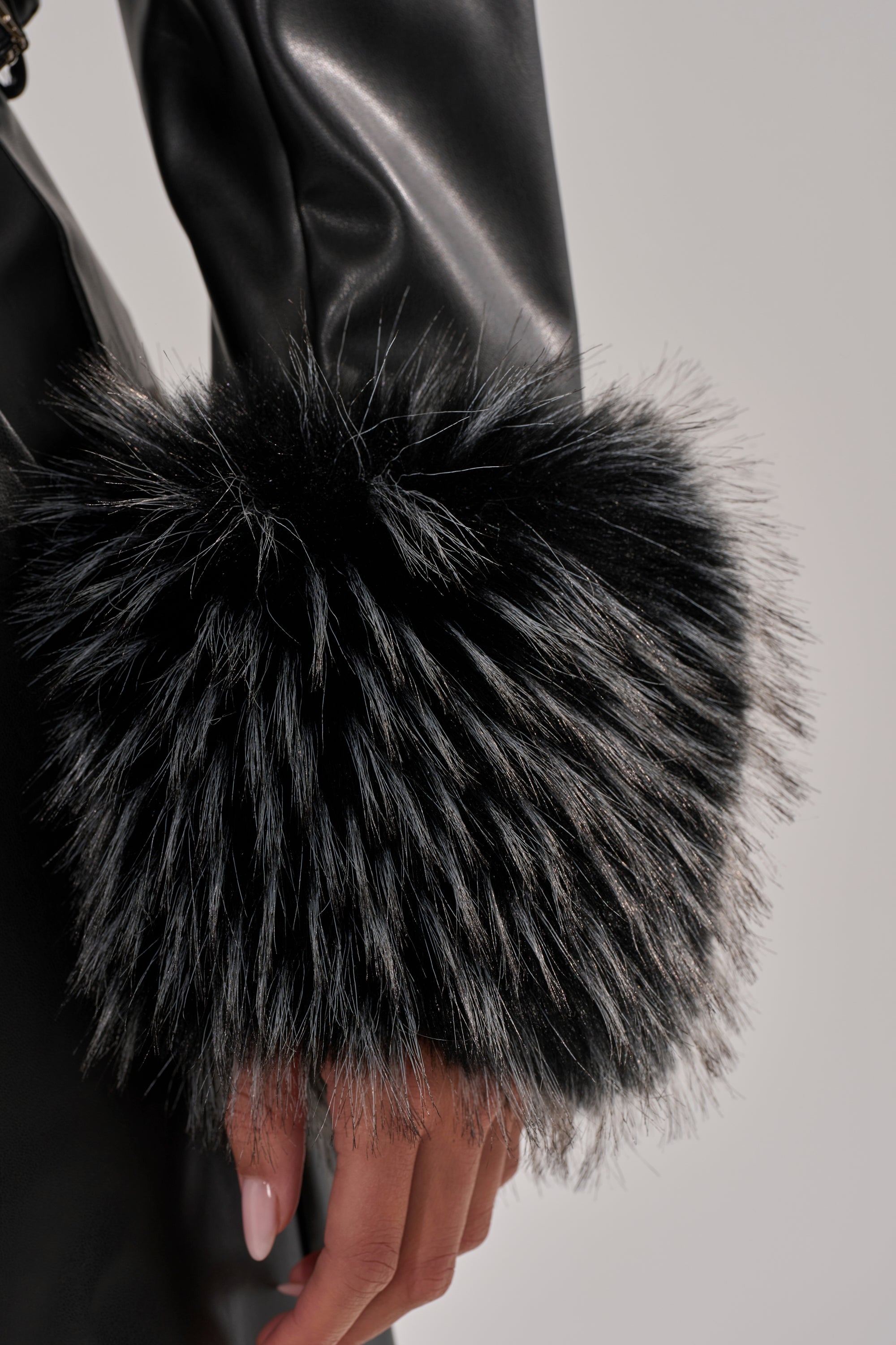 A close-up of a person’s arm wearing the MIND YOUR OWN FUR TRIM BLAZER, featuring black faux leather and a large, fluffy black and white faux fur cuff; their hand rests at their side.