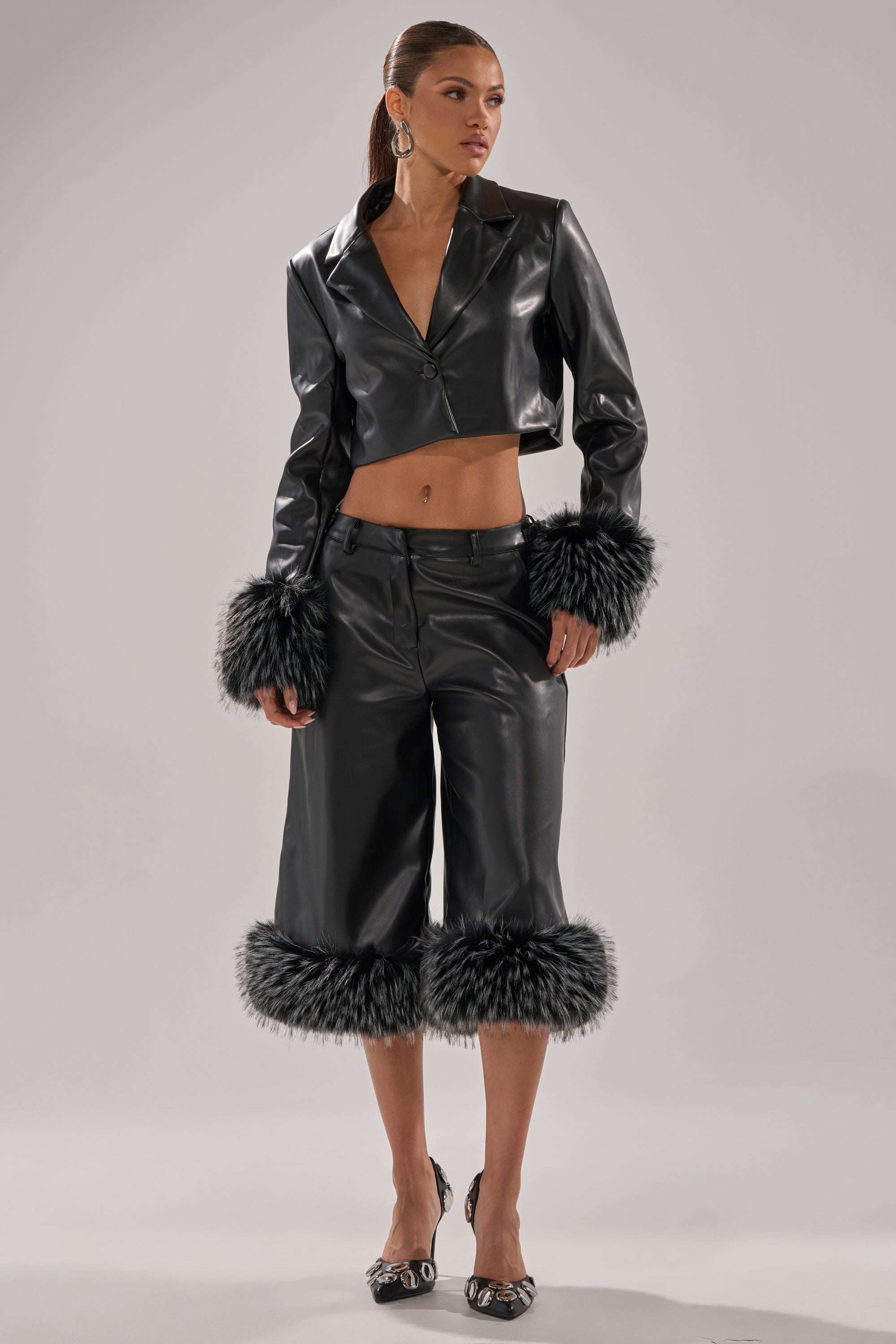A woman wears the MIND YOUR OWN FUR TRIM BLAZER, a black cropped faux leather piece with black-and-gray faux fur cuffs, paired with matching culottes. She completes the look with hoop earrings, pointed heels, and tied-back hair.