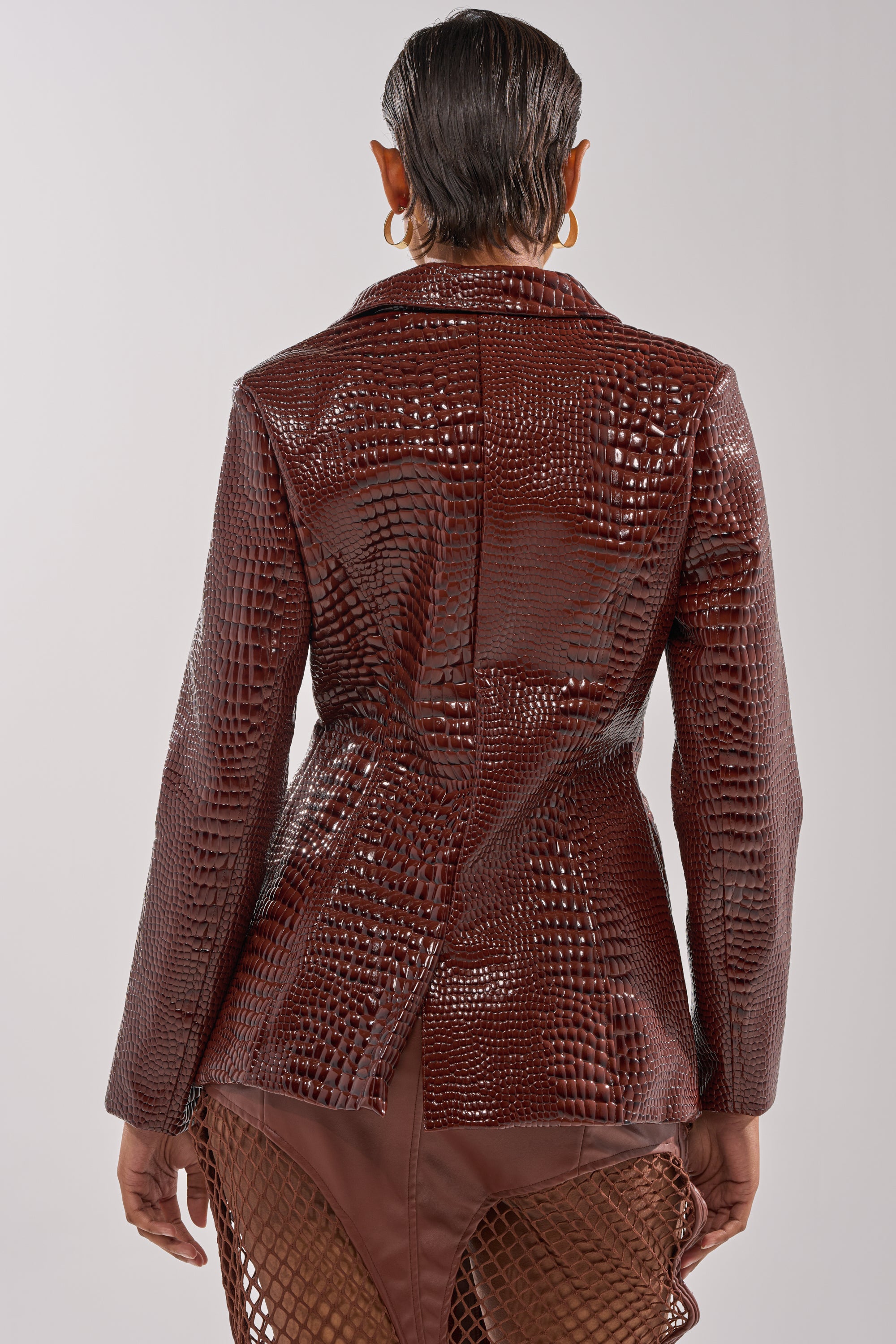 A person with short, slicked-back hair is shown from the back in the NEW FLAME EMBOSSED PATENT BLAZER, a shiny brown crocodile-patterned faux patent piece with a matching mesh-detailed skirt, set against a plain light background. 4