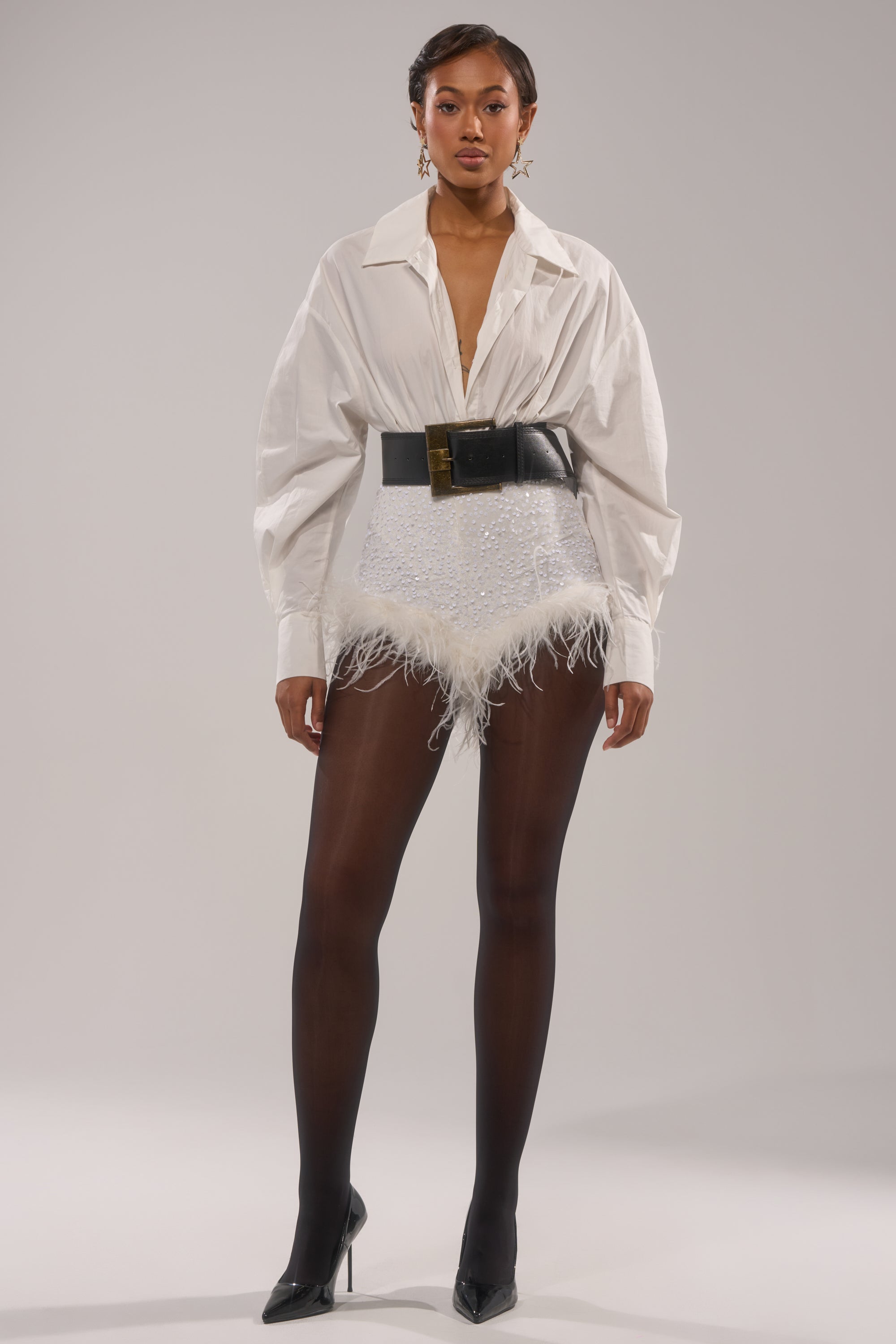 A woman exudes confidence in white oversized button-down, high-waisted NEW YEAR FEATHER SHORTS with sequins, a wide black belt, sheer tights, and black pointed heels. 8