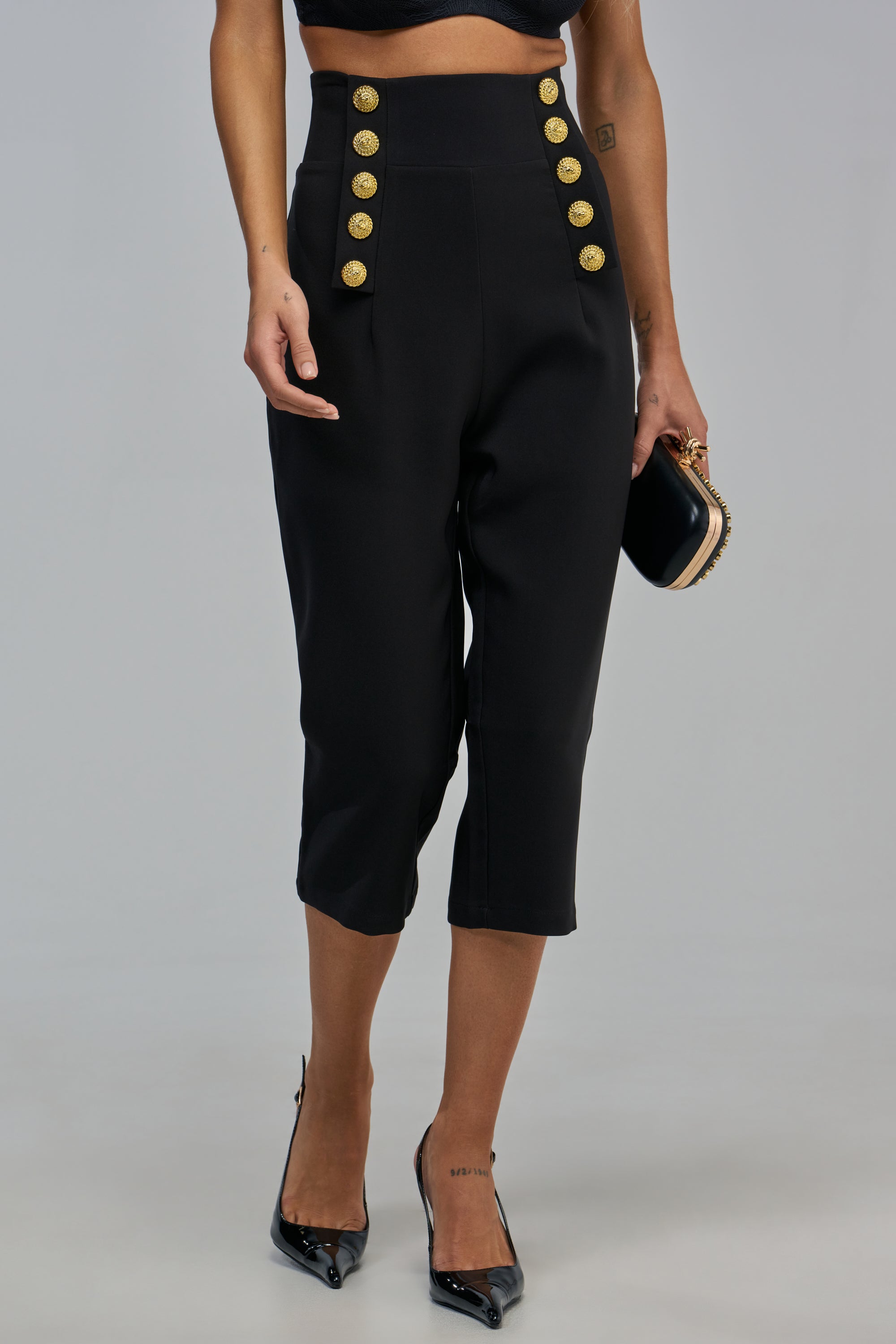 CALL ME CUTE CAPRI high-waisted black capri pants featuring two columns of decorative gold buttons, worn with black pointed-toe heels and a small black clutch for a sleek, business casual look.