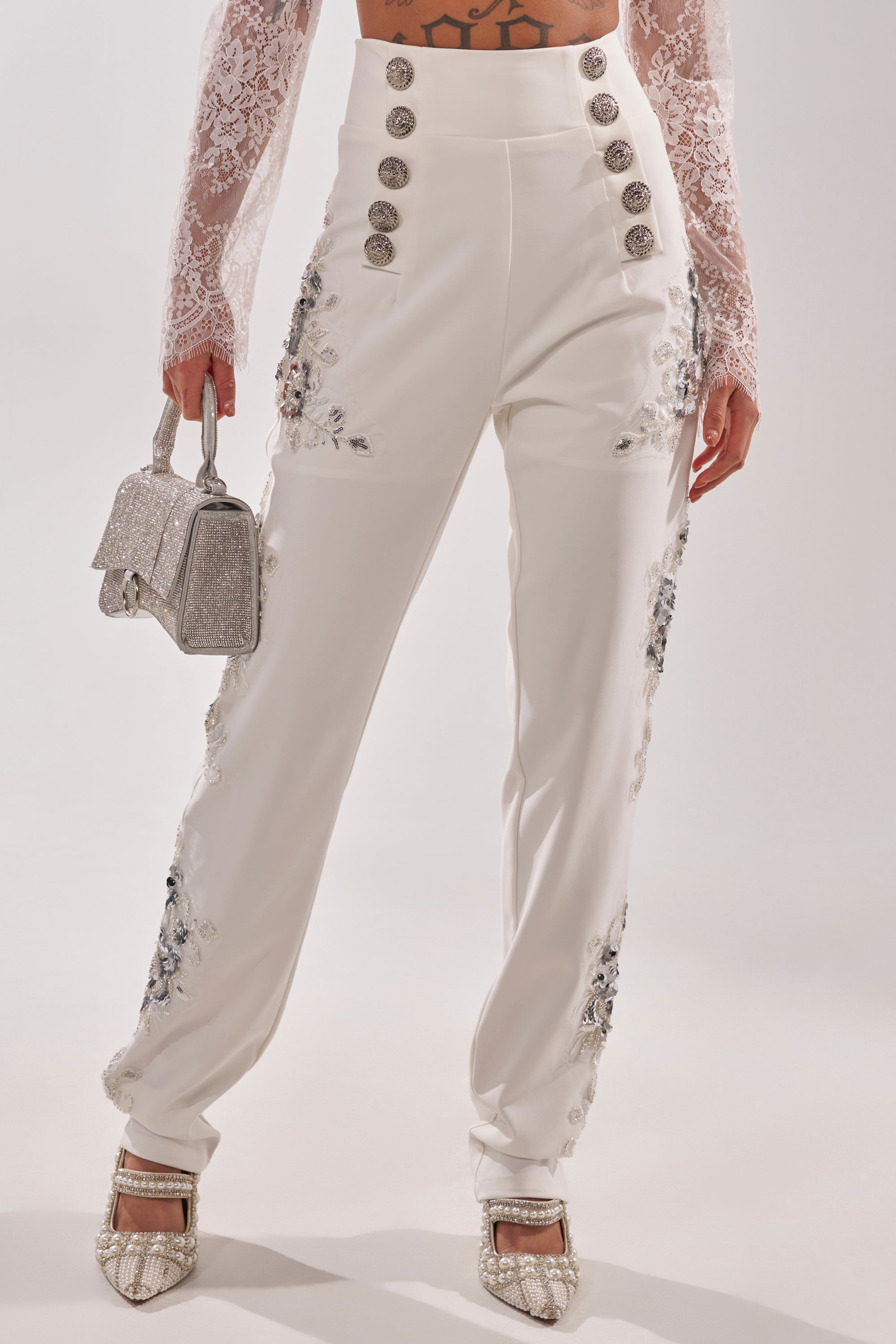 Wearing the VENETIAN EMBELLISHED TROUSER—high-waisted white trousers with decorative buttons and silver floral embroidery—a lace long-sleeved crop top, glittery silver handbag, and crystal-embellished heels. 0