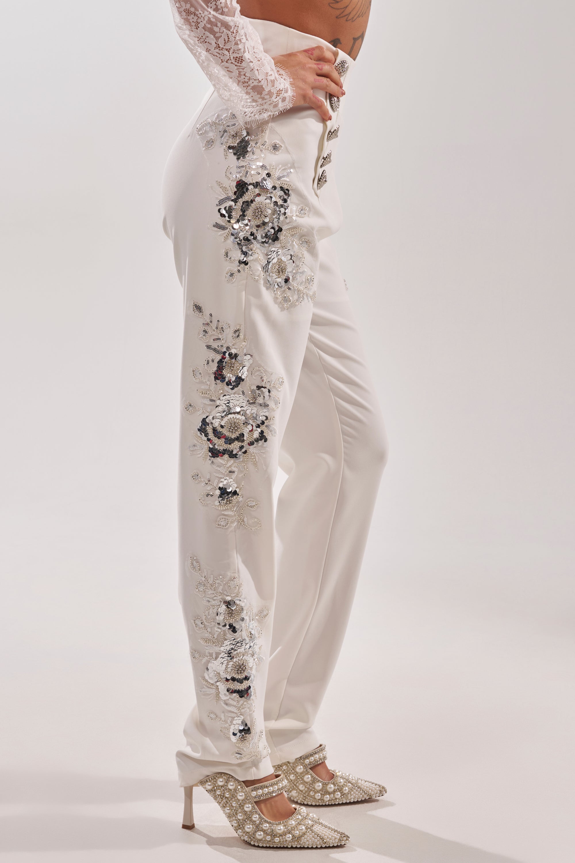 A person stands sideways against a plain white background, wearing the VENETIAN EMBELLISHED TROUSER featuring silver sequin floral and crystal embroidery, paired with laced white high heels. 1