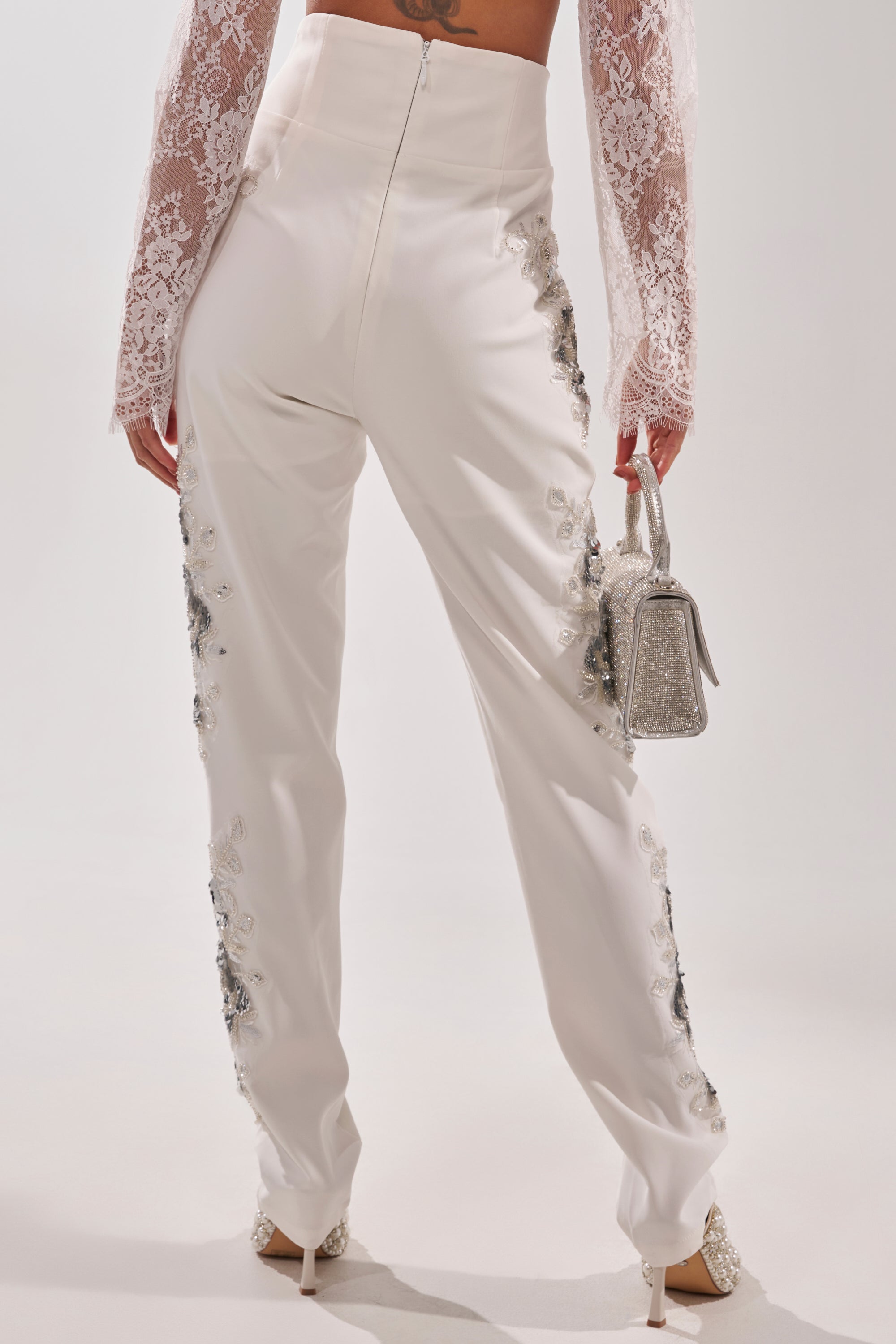 Wearing the VENETIAN EMBELLISHED TROUSER—high-waisted, white with crystal embroidery—a person pairs it with lace sleeves, a small glittery silver handbag, and white heels, standing facing away. 2