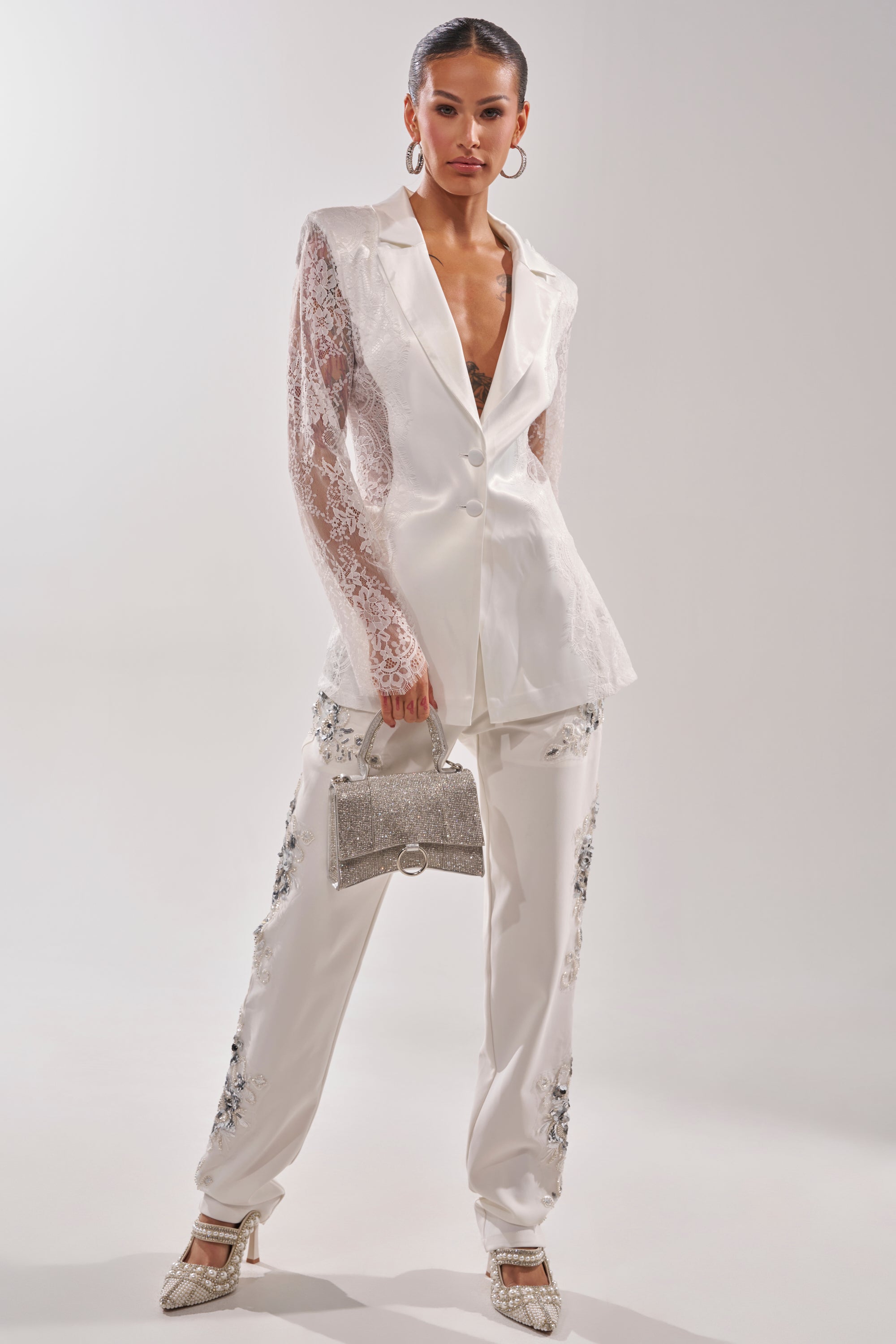 A woman stands confidently in a white suit featuring the VENETIAN EMBELLISHED TROUSER with lace sleeves, holding a sparkling silver handbag. She completes her look with high heels, hoop earrings, and her hair pulled back. 3