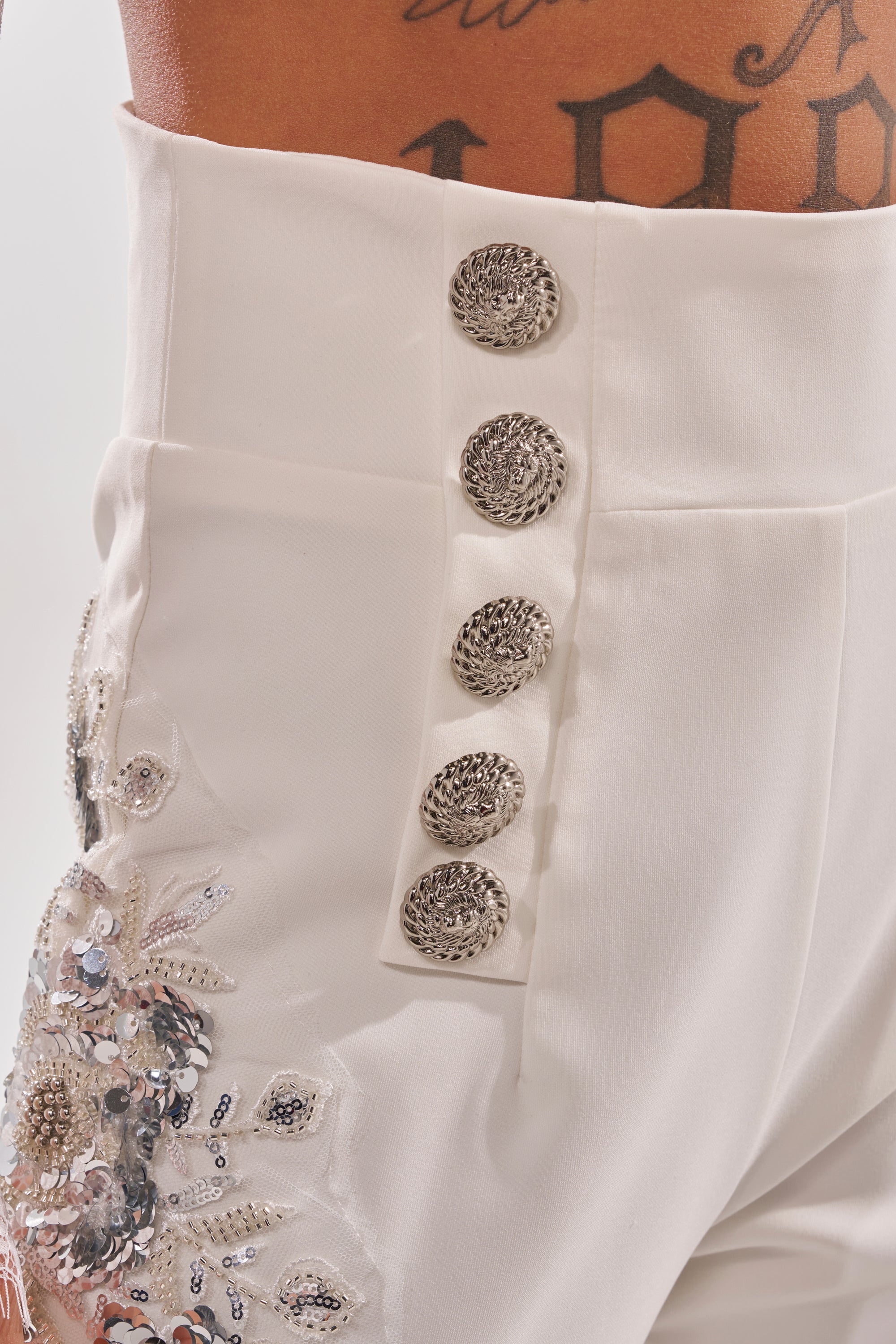 Close-up of the VENETIAN EMBELLISHED TROUSER in white, featuring five ornate silver buttons, floral sequin and crystal embroidery on the hip, plus a tattoo visible on the wearer’s lower abdomen. 5