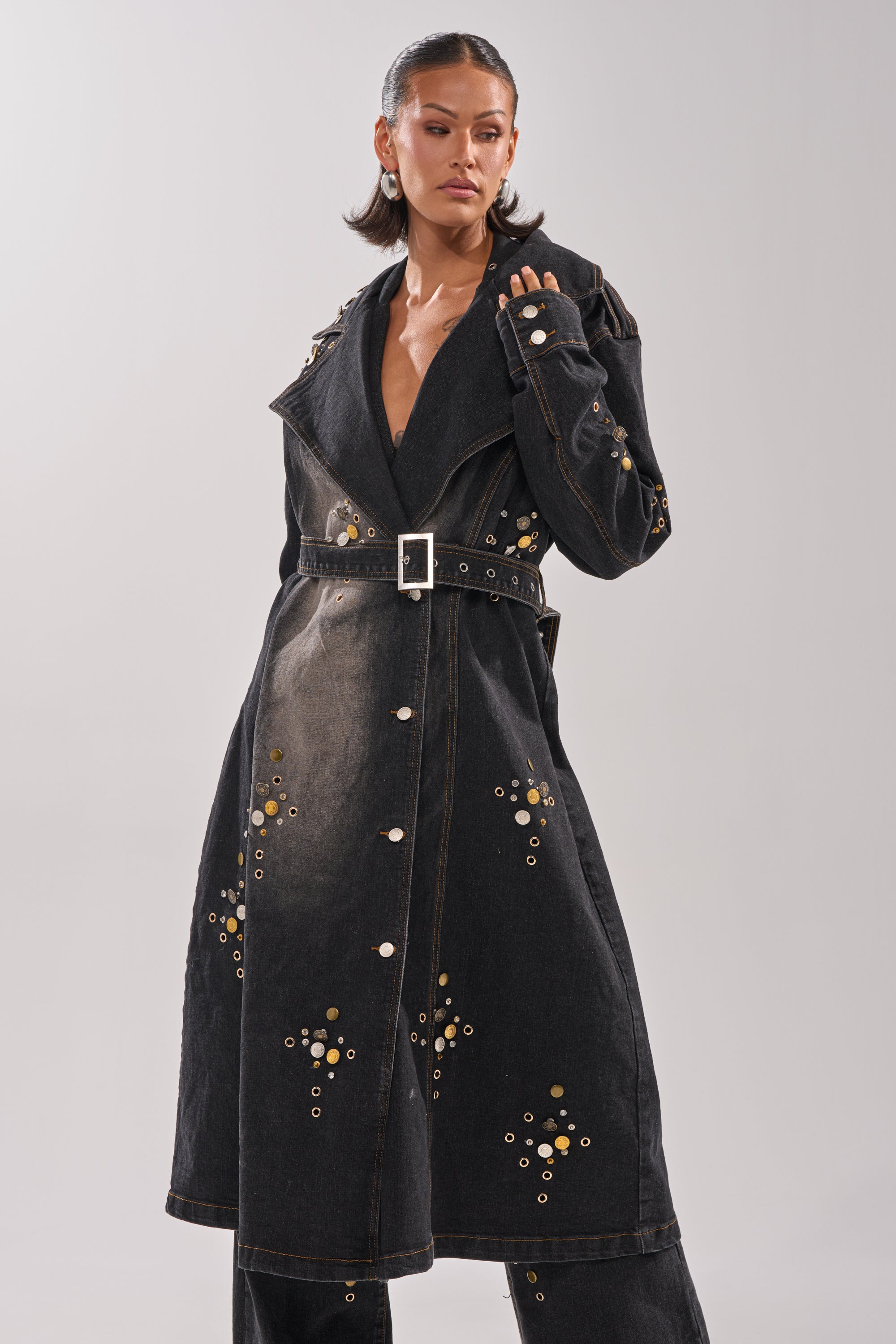 A woman stands confidently against a plain light background wearing the SPACE AND TIME STUDDED DENIM TRENCH, featuring metallic embellishments and a belted waist.
