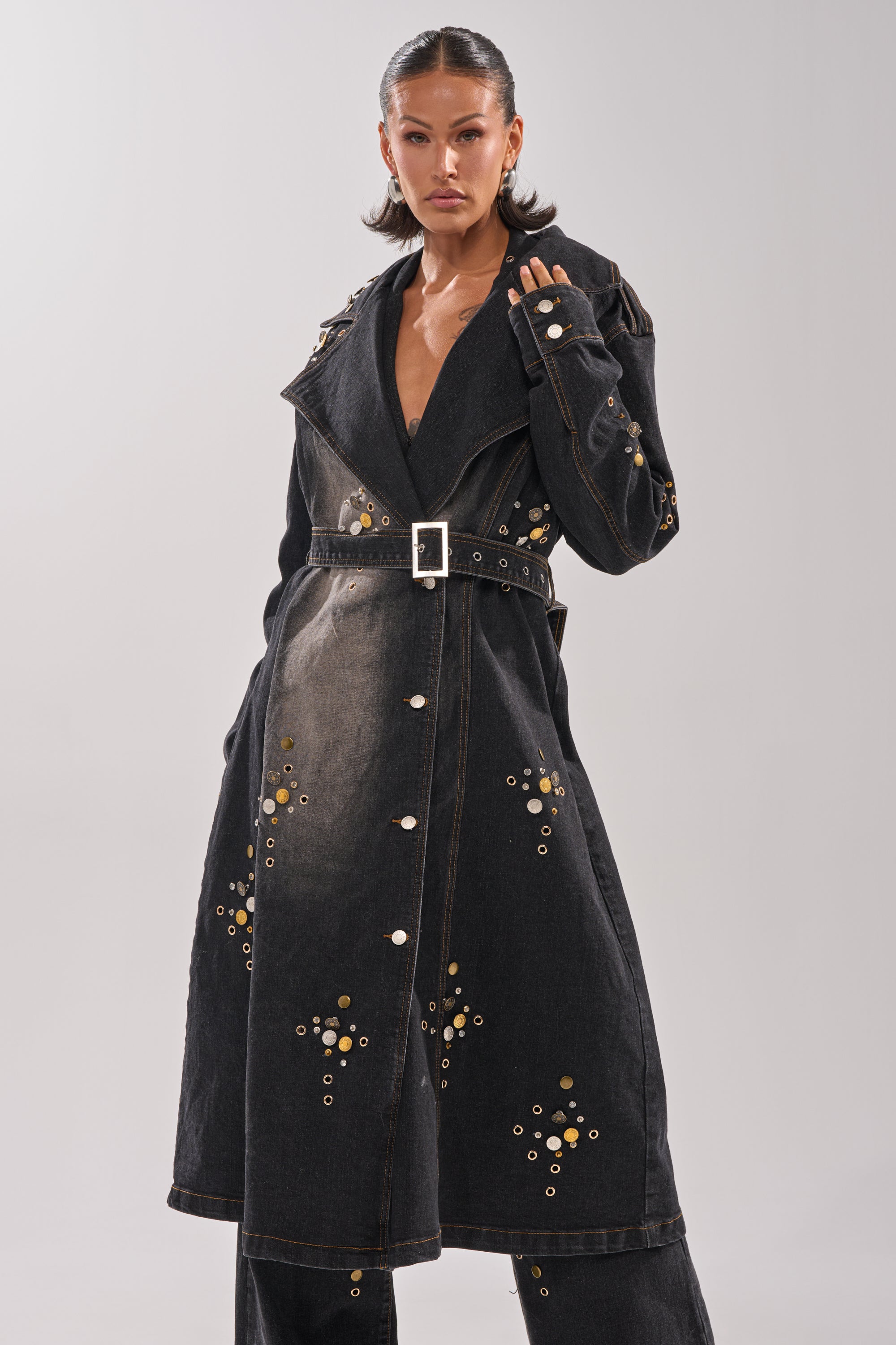 A woman stands confidently in the SPACE AND TIME STUDDED DENIM TRENCH, a longline studded denim coat with a wide collar, belted waist, metallic circular embellishments, silver snaps, and matching dark denim pants.