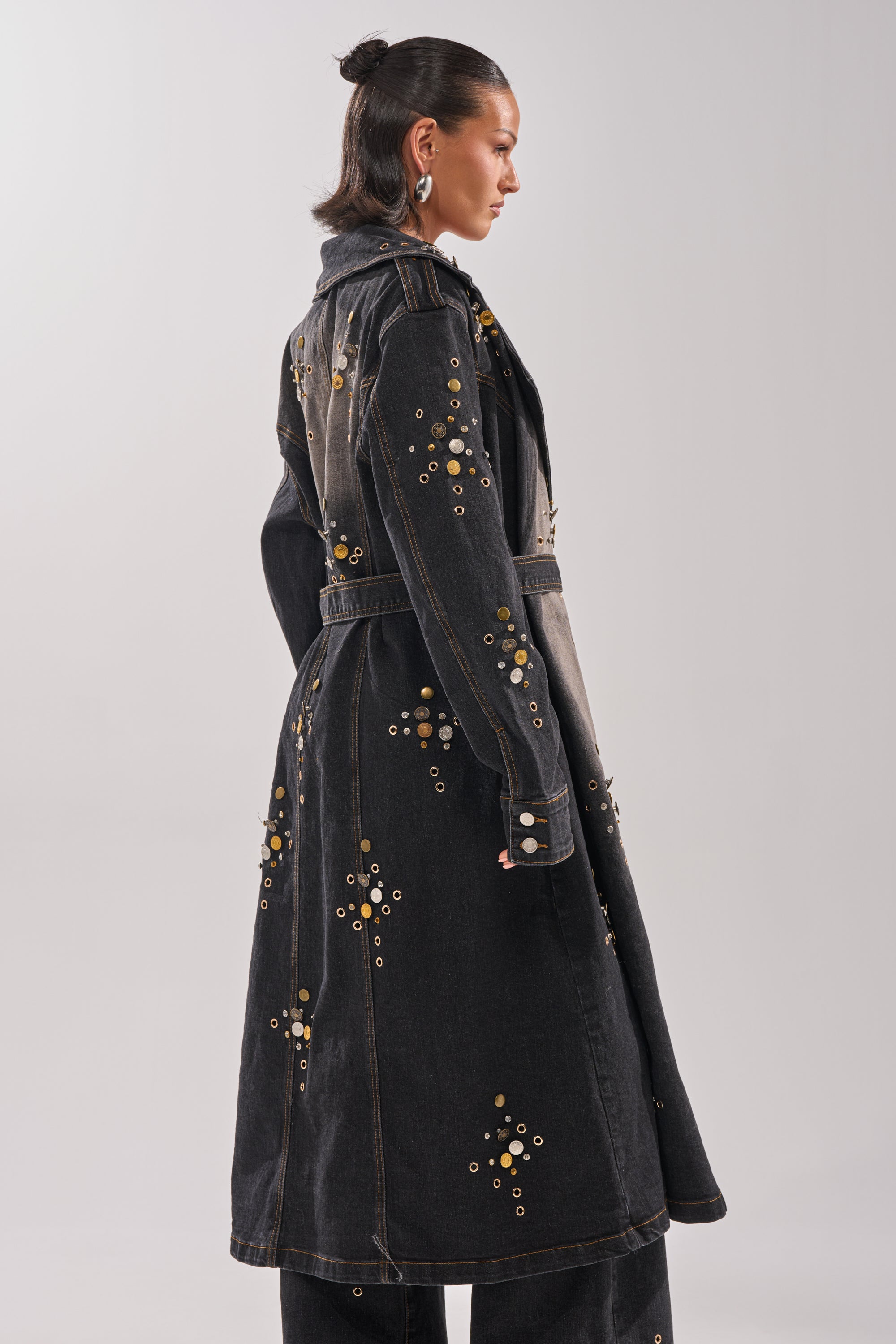 A woman in profile wears the SPACE AND TIME STUDDED DENIM TRENCH, featuring gold grommets, a belted waist, and longline silhouette, paired with matching wide-leg pants, slicked-back hair, and statement earrings.