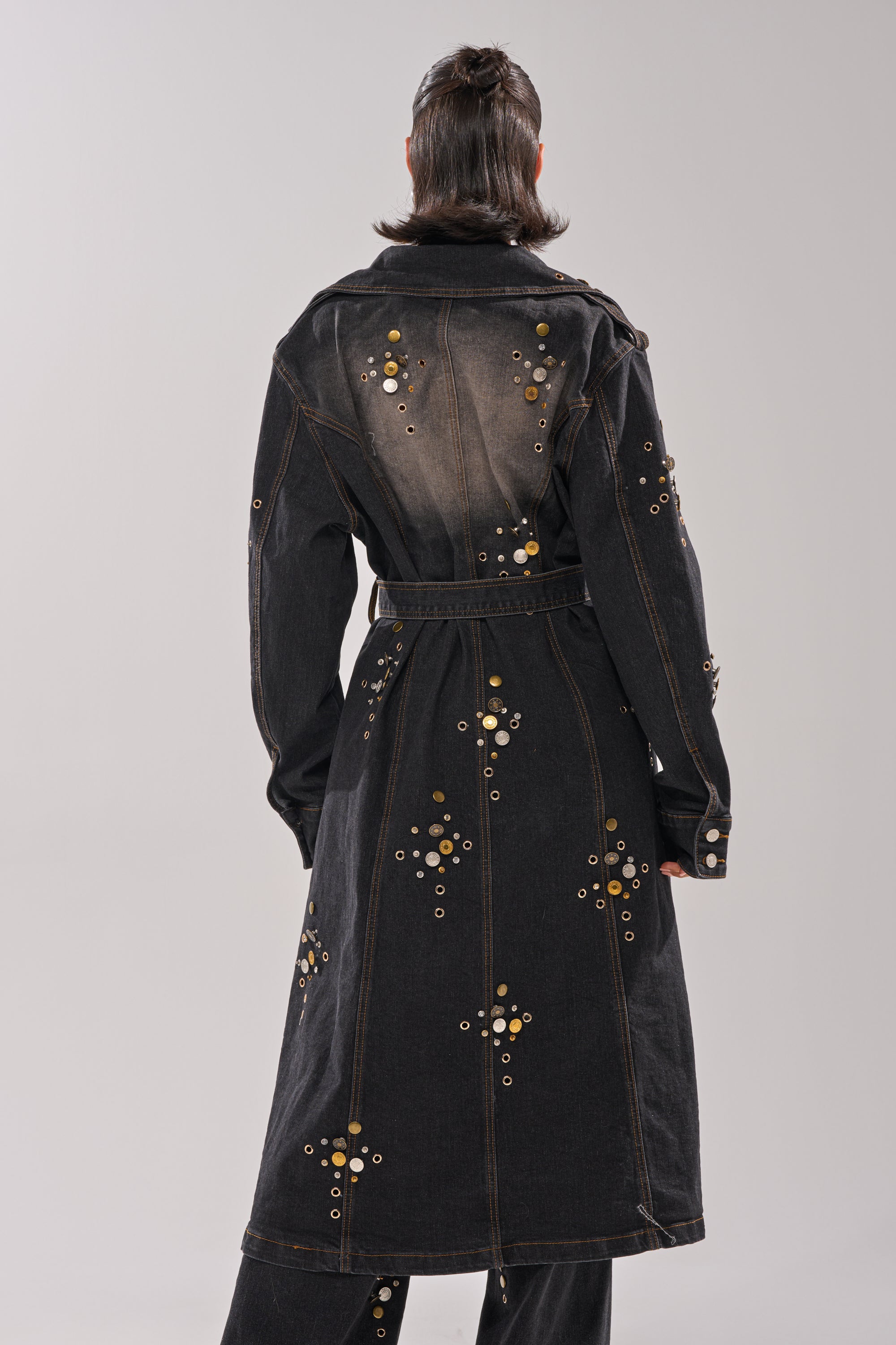 A person faces away, wearing the SPACE AND TIME STUDDED DENIM TRENCH with metallic eyelets, grommets, and a belted waist over black pants. The long coat stands out against a plain light background.