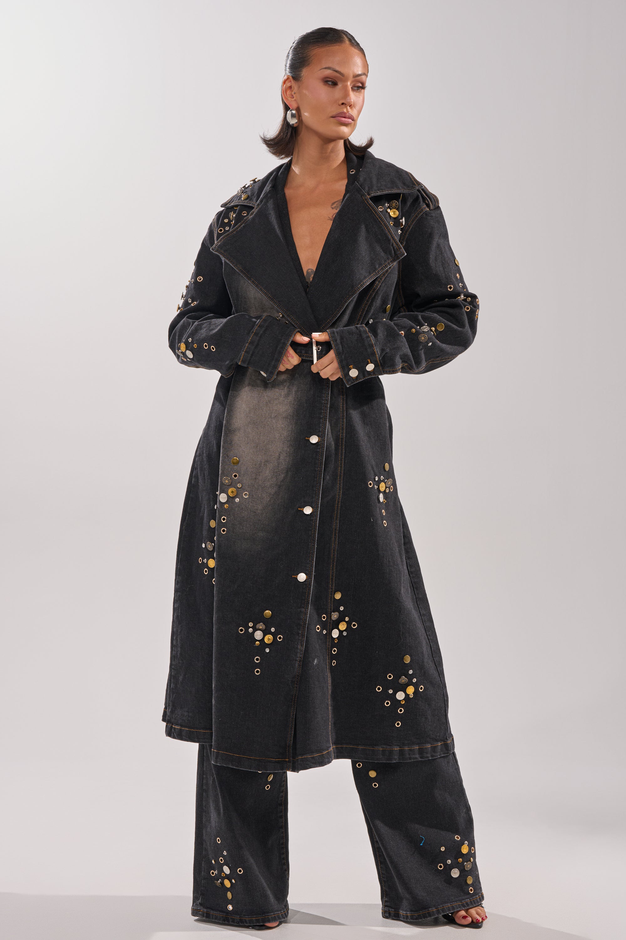 A woman stands confidently in the SPACE AND TIME STUDDED DENIM TRENCH with metallic studs, matching wide-leg pants, hoop earrings, and her hair sleekly pulled back against a plain background.
