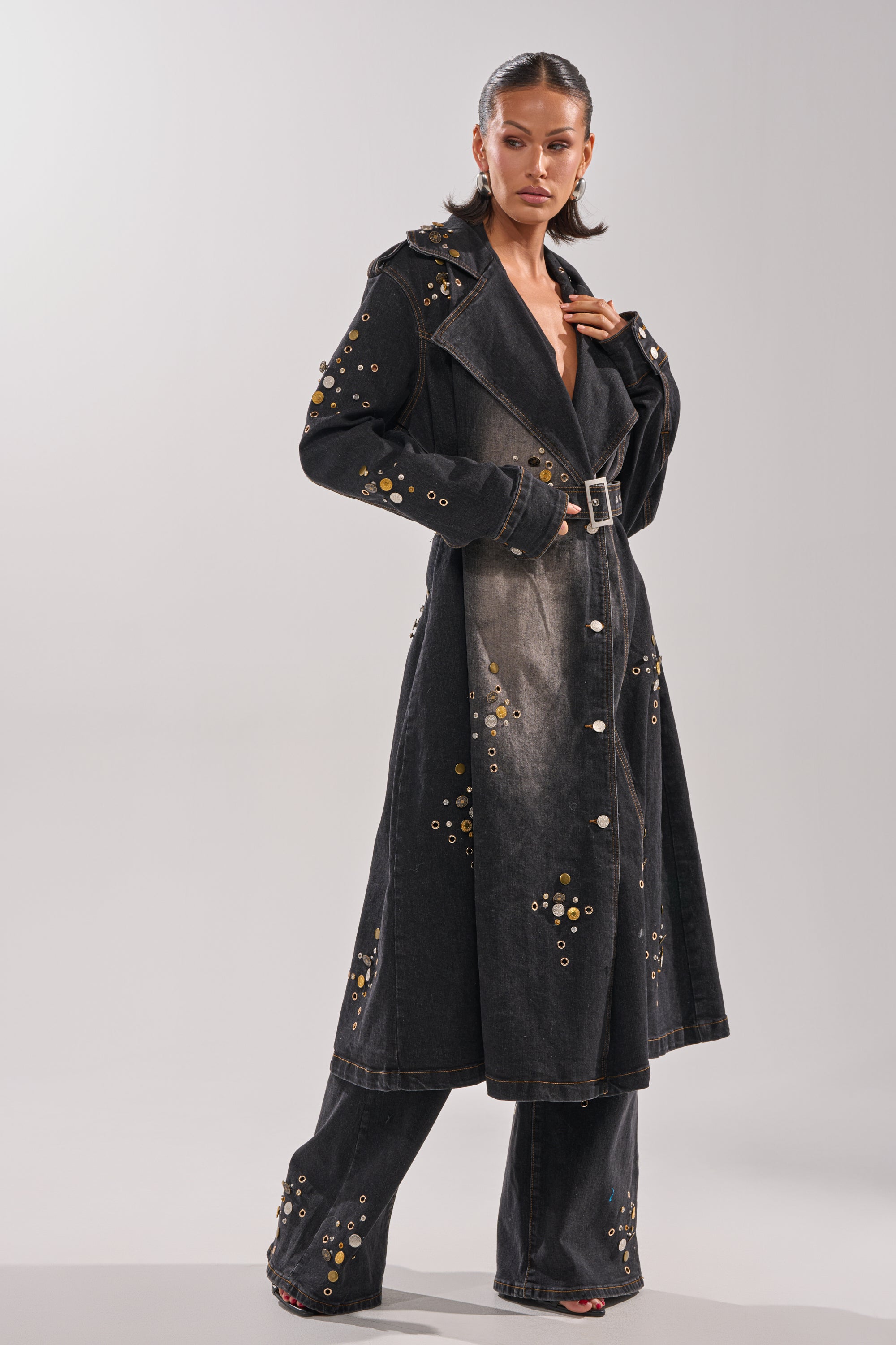 A woman models the SPACE AND TIME STUDDED DENIM TRENCH, featuring metallic eyelet details, paired with wide-leg matching pants against a simple light background.