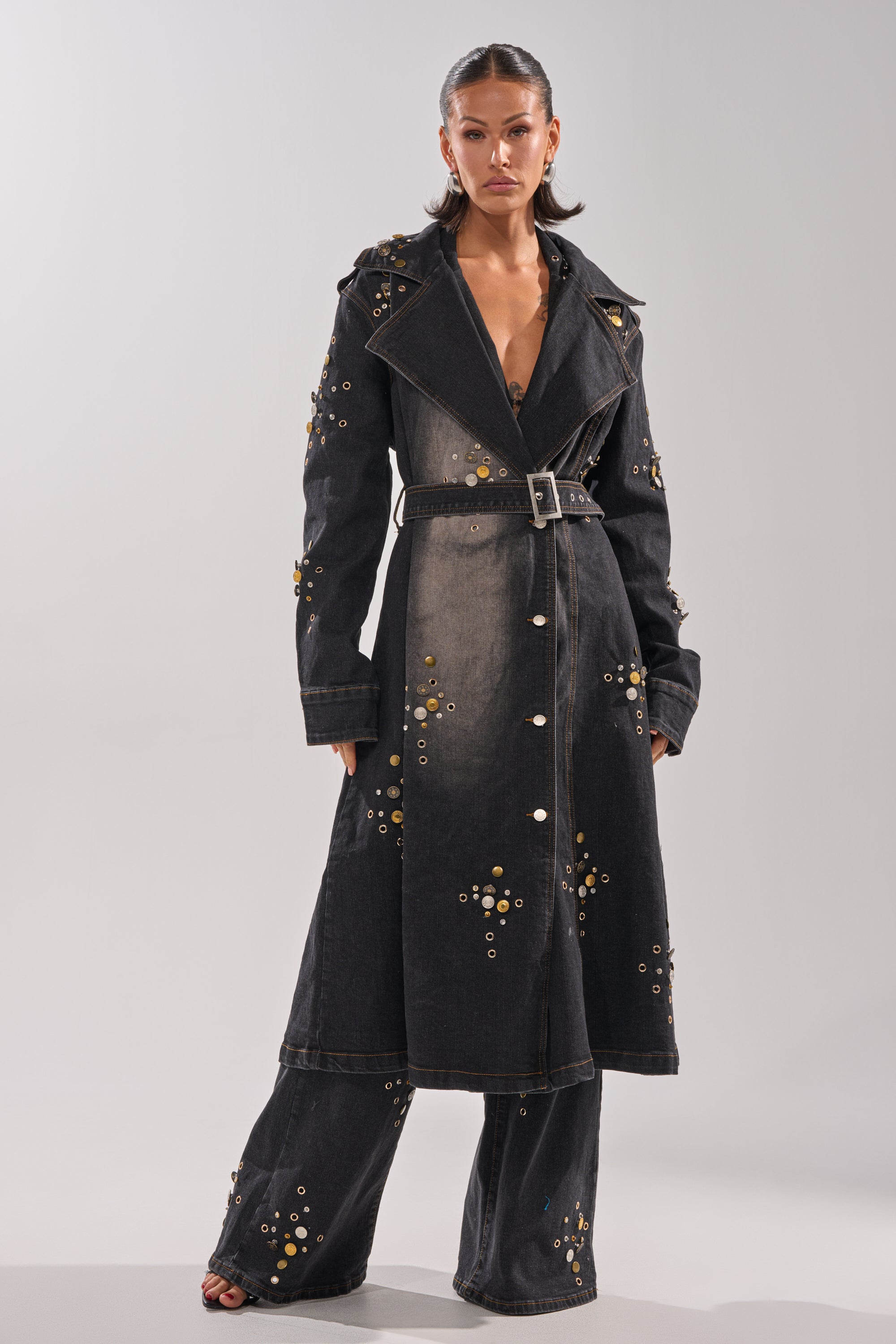 A woman with slicked-back hair and a confident expression models the SPACE AND TIME STUDDED DENIM TRENCH and matching wide-leg pants, both detailed with metallic grommets, against a simple backdrop.