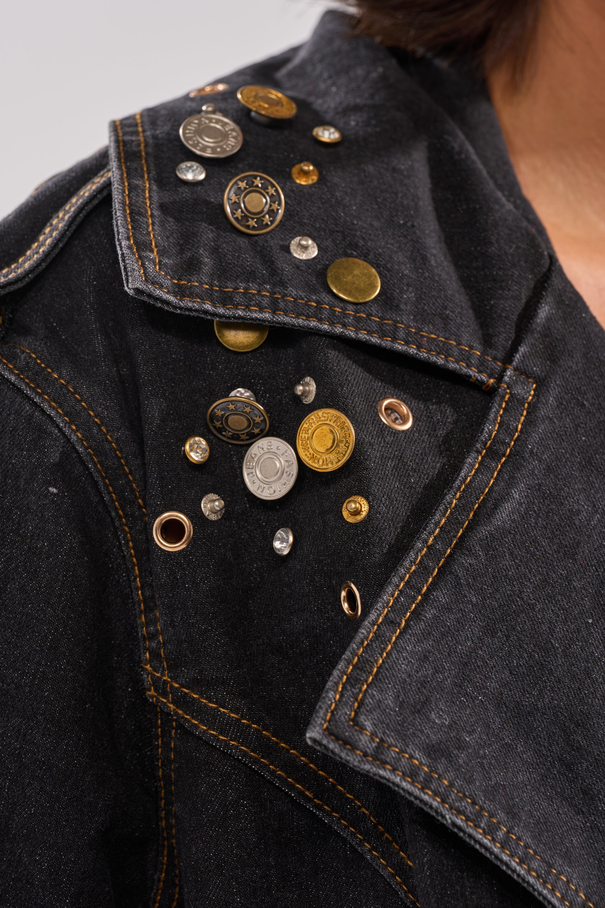 A close-up of the SPACE AND TIME STUDDED DENIM TRENCH, a black denim jacket with orange stitching, featuring assorted metal buttons, studs, and eyelets on the lapel near the shoulder.