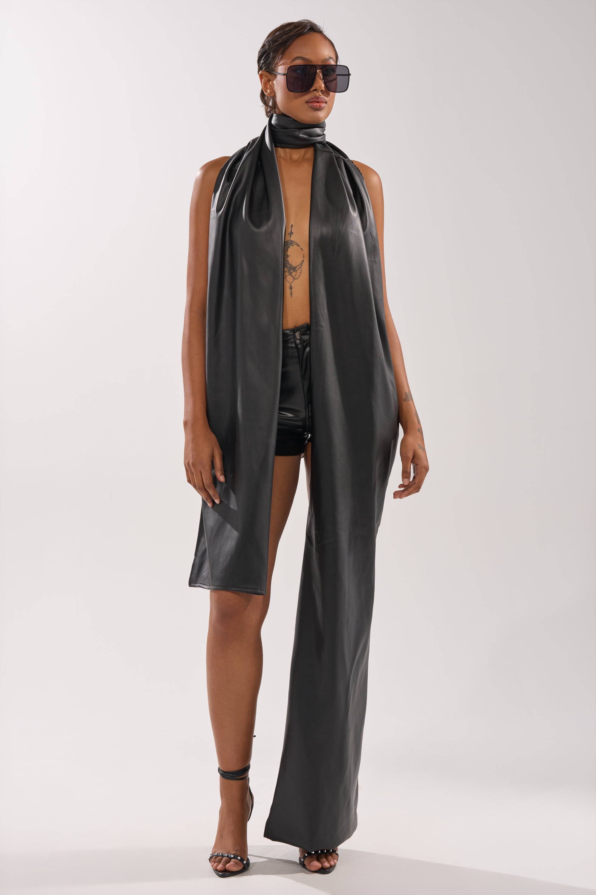 A person models the VALLY PU SCARF PANT: a sleeveless, asymmetrical black outfit with one pant leg and matching faux leather scarf, styled with large sunglasses and high heels, posing confidently against a plain background. 2