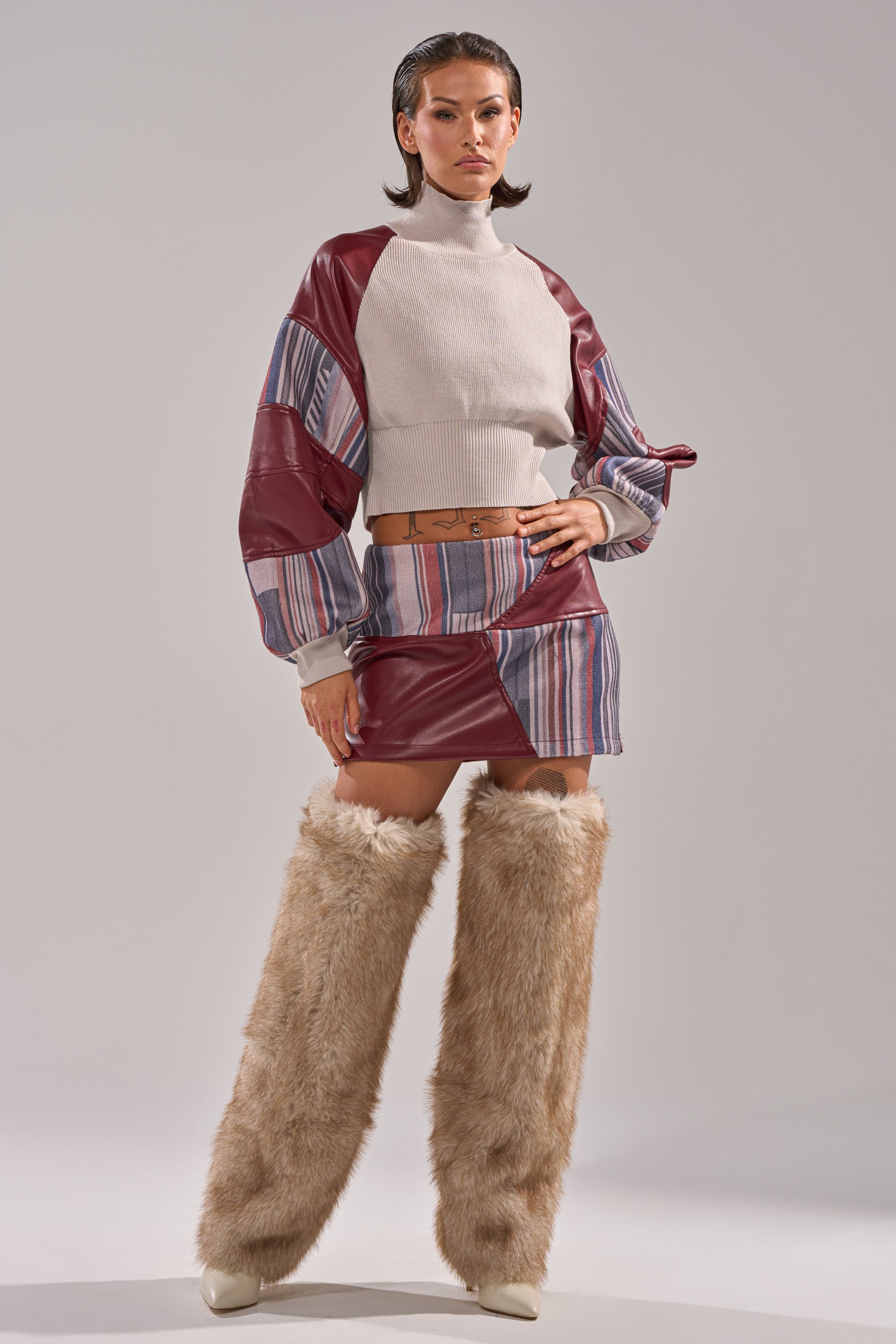 A woman stands confidently in the FEELING THE VIBES MINI SKIRT, paired with a beige patchwork turtleneck, maroon faux leather sleeves, a tan belt, and tall furry beige leg warmers over pointed shoes against a plain background.