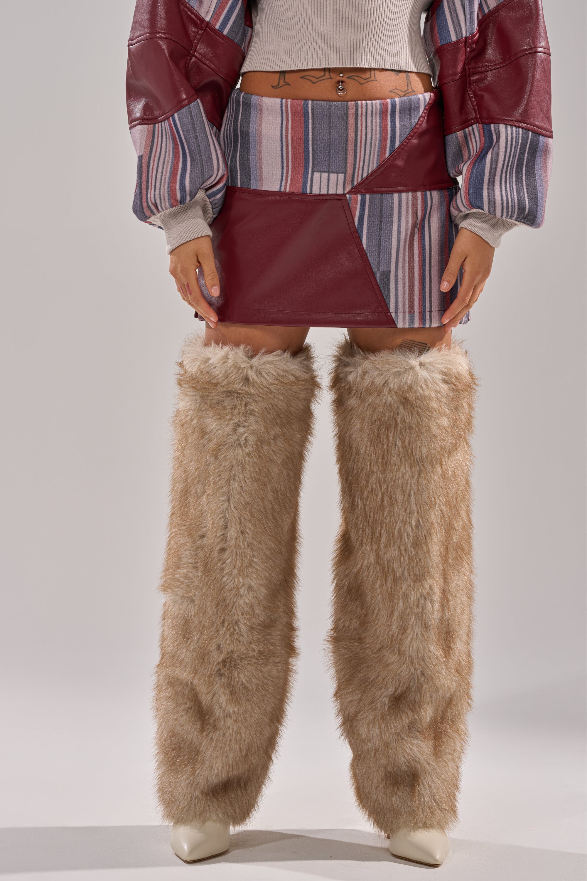 Wearing the FEELING THE VIBES MINI SKIRT, a person pairs it with a cropped jacket, midriff-baring top showing a stomach tattoo, beige furry leg warmers, and white heeled boots.