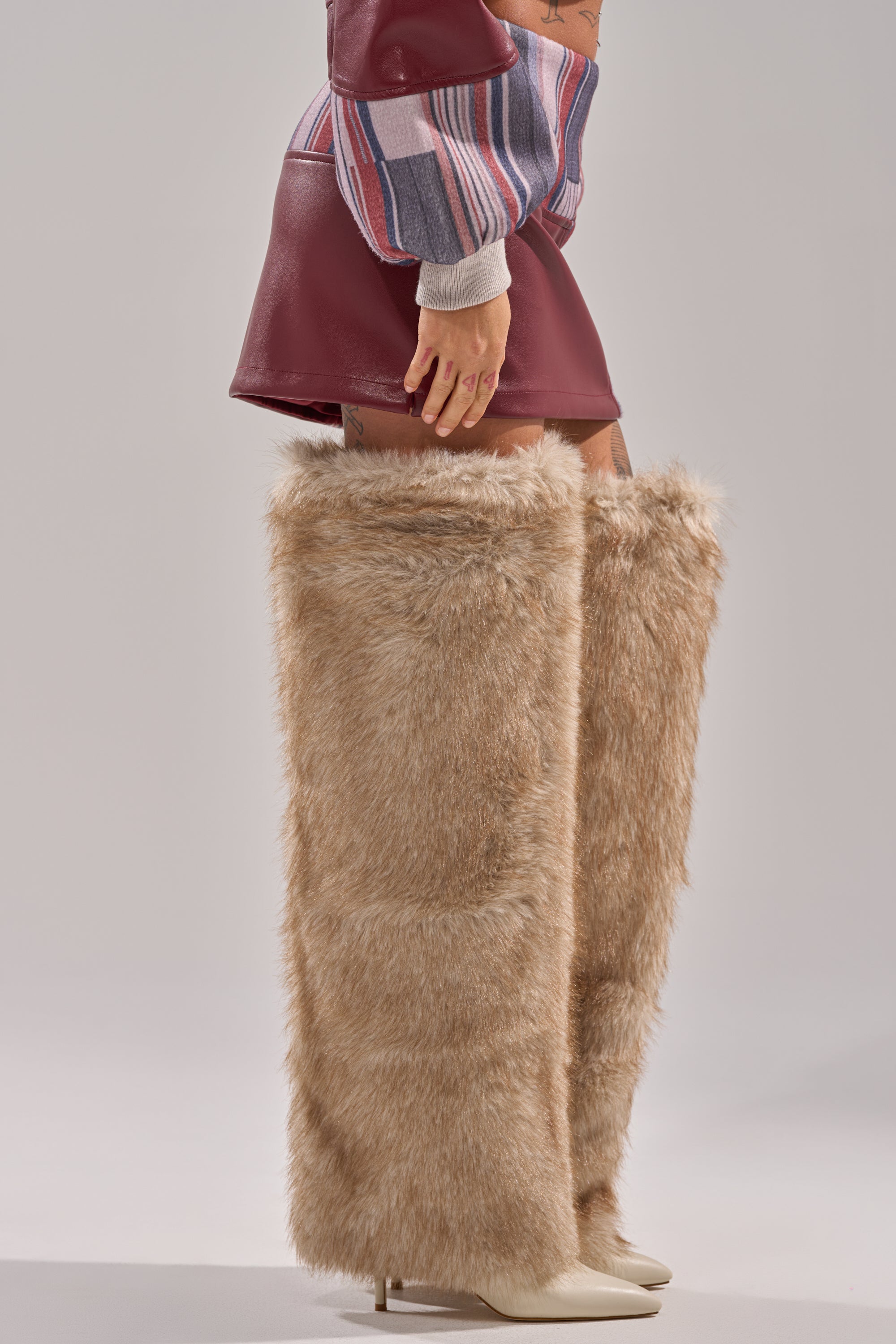 A person wearing the FEELING THE VIBES MINI SKIRT, a colorful patchwork long-sleeve top, and tall light brown faux fur boots with pointed white heels visible beneath.