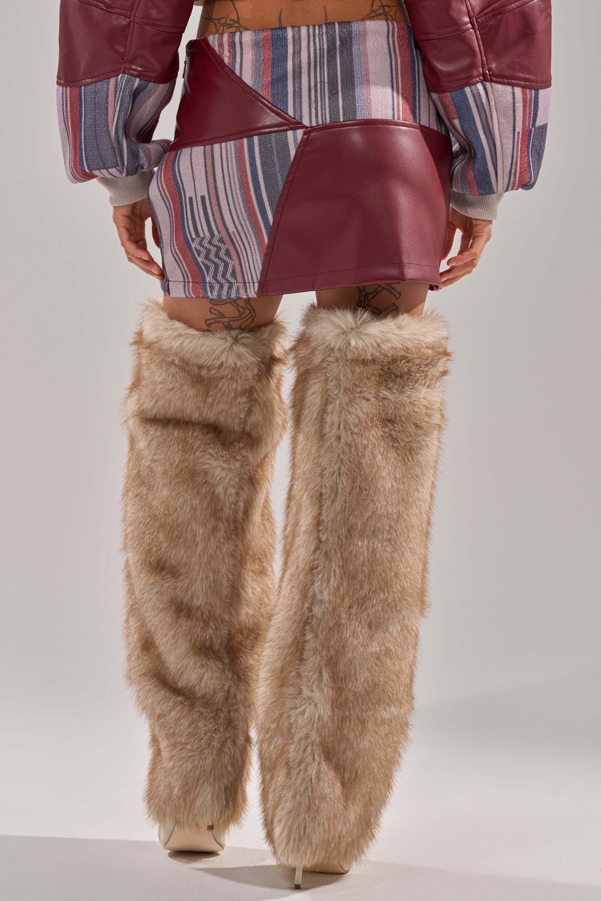A person is seen from behind wearing the FEELING THE VIBES MINI SKIRT, featuring red faux leather and patterned panels, paired with oversized beige furry leg warmers against a neutral background.