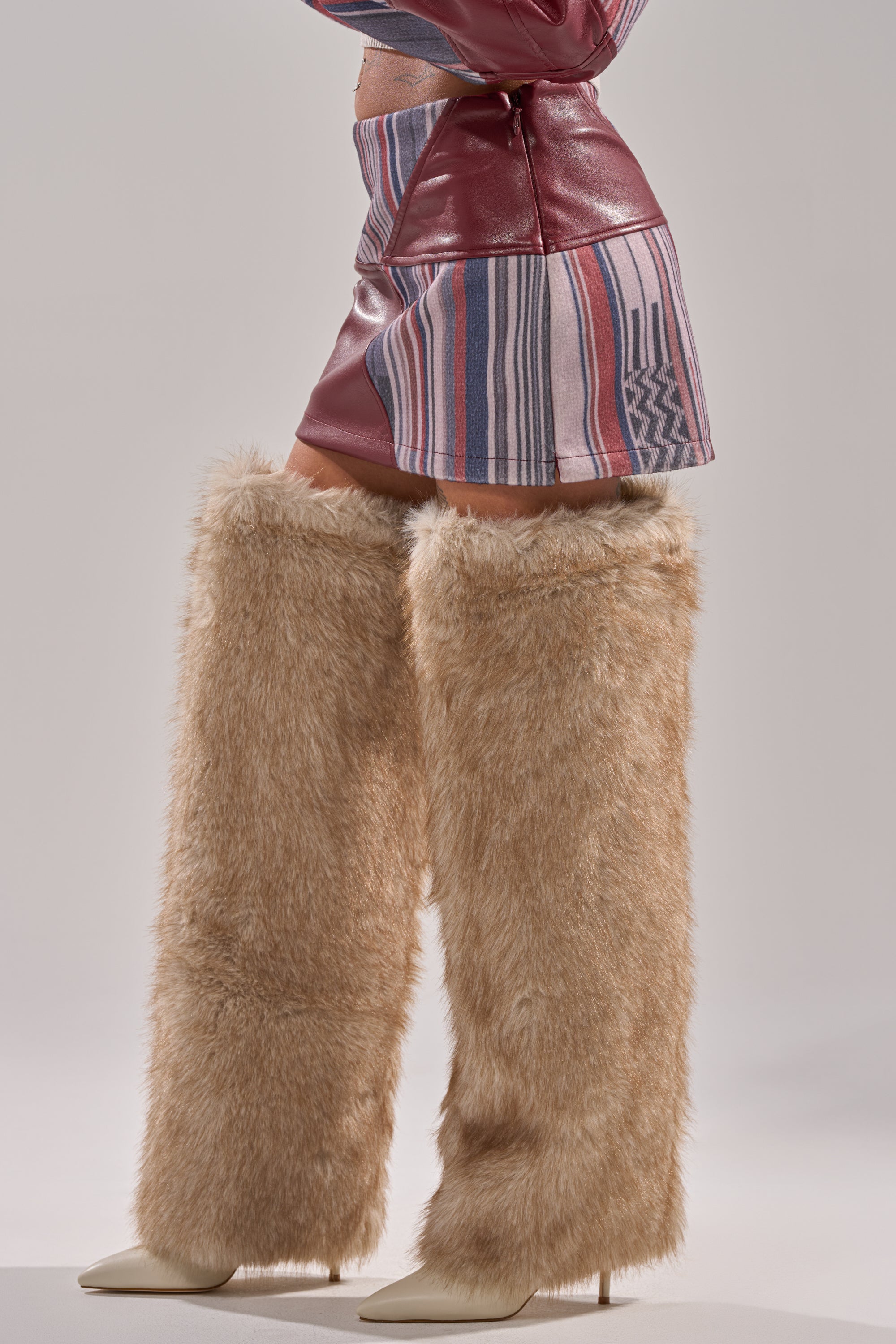 A person wearing the FEELING THE VIBES MINI SKIRT, a maroon jacket, and tall furry tan leg warmers over white pointed-toe heels, stands against a neutral background.