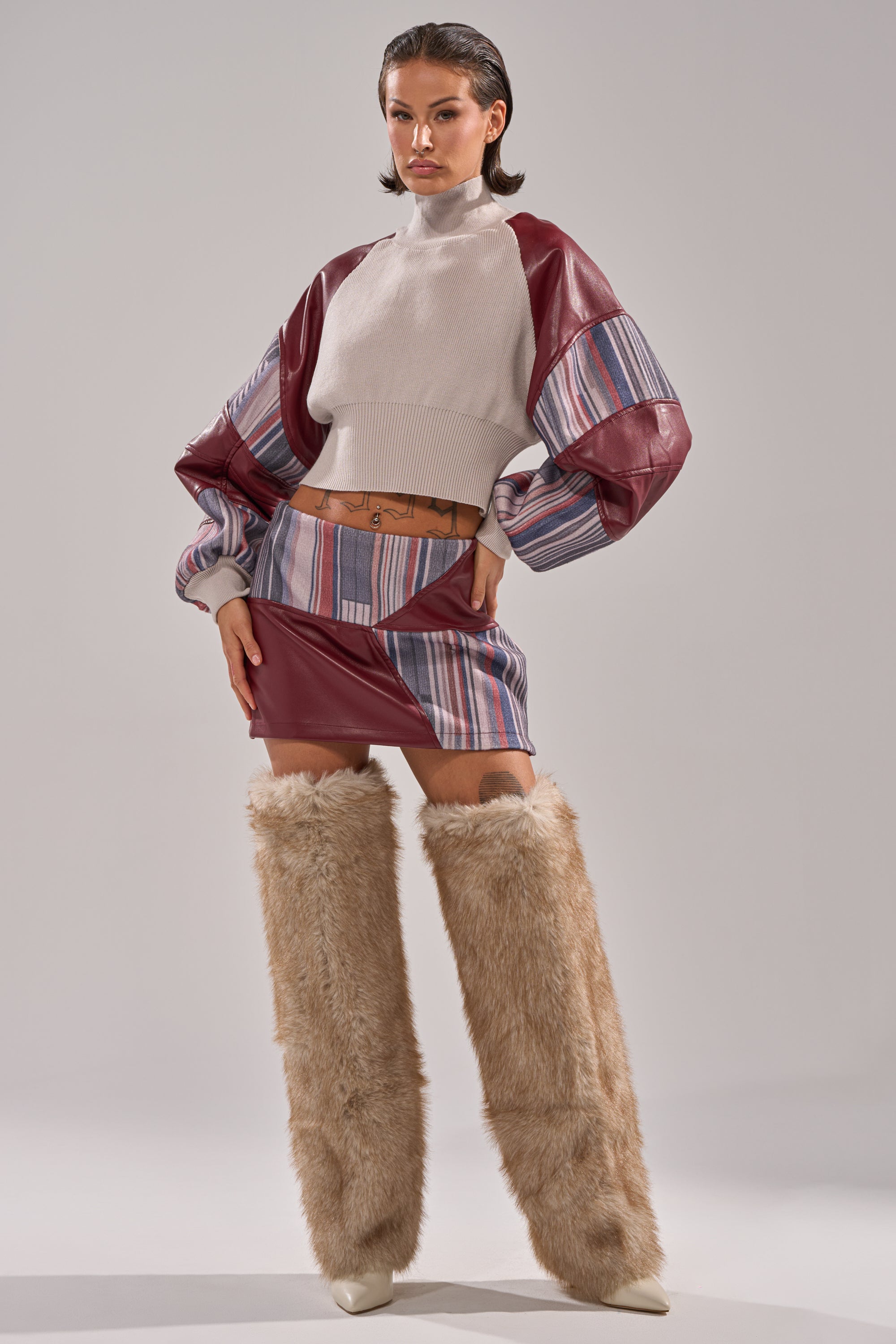 A woman exudes chic style in a high-neck cream sweater with burgundy striped puffy sleeves, the FEELING THE VIBES MINI SKIRT, a tan belt, and tall furry beige boots against a plain background.