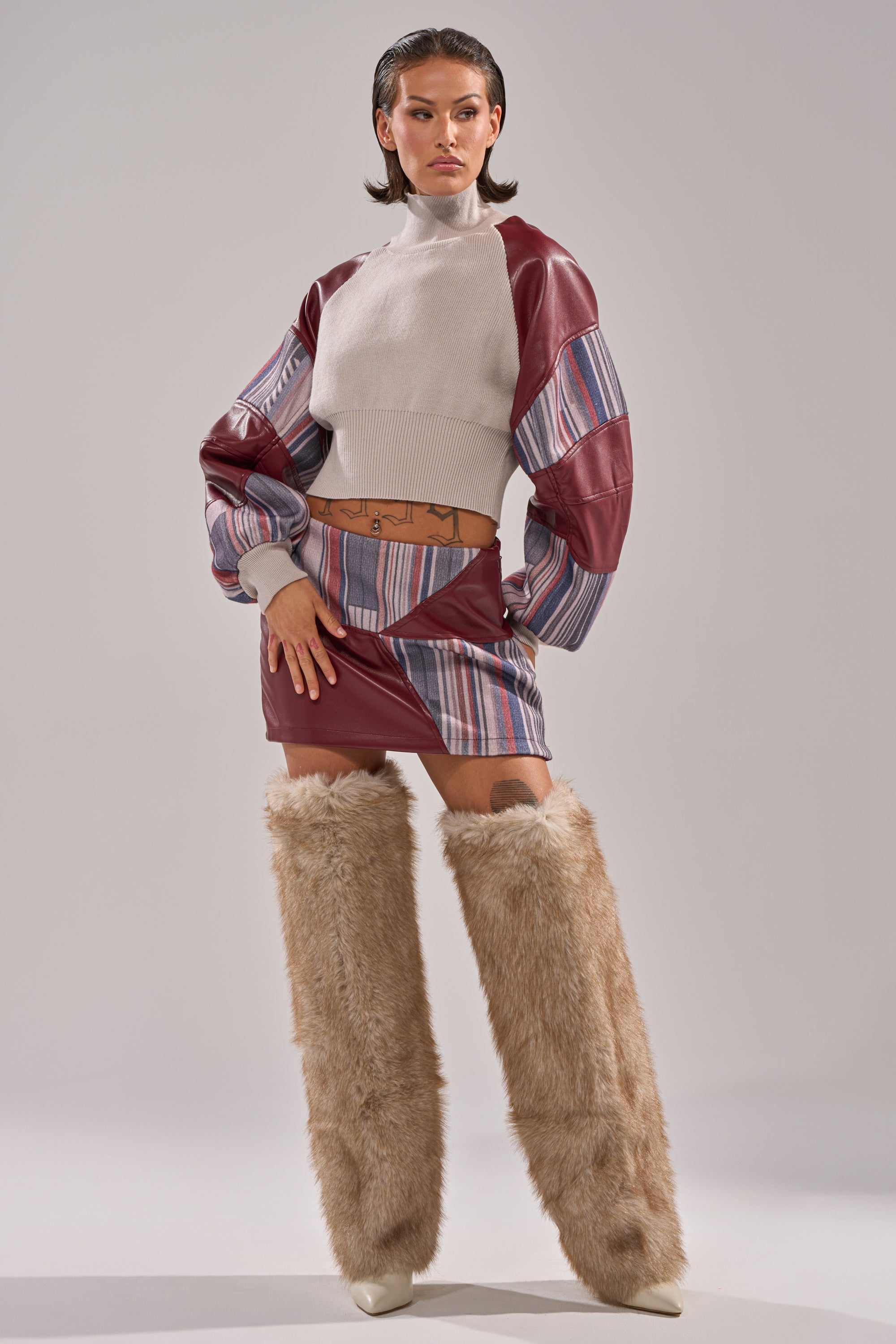 A woman poses confidently in a contemporary style, wearing the FEELING THE VIBES MINI SKIRT, a white turtleneck crop top, matching sleeves, and tall light brown furry boots against a plain light gray background.