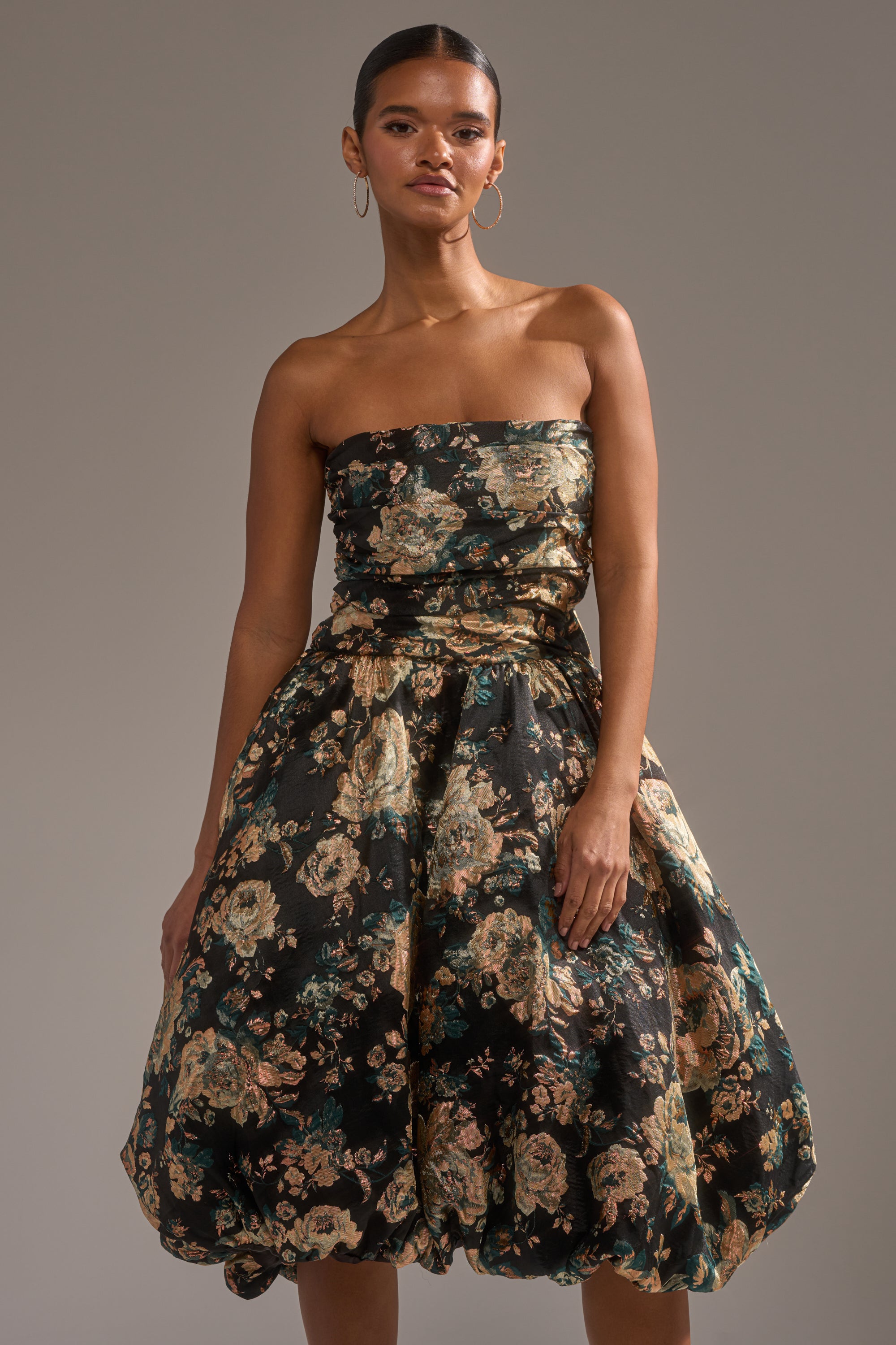 A woman wears the DRESSED TO IMPRESS BROCADE MIDI DRESS, a strapless evening dress with a voluminous gold and green floral metallic skirt, standing against a neutral background with slicked-back hair and large hoop earrings. 1