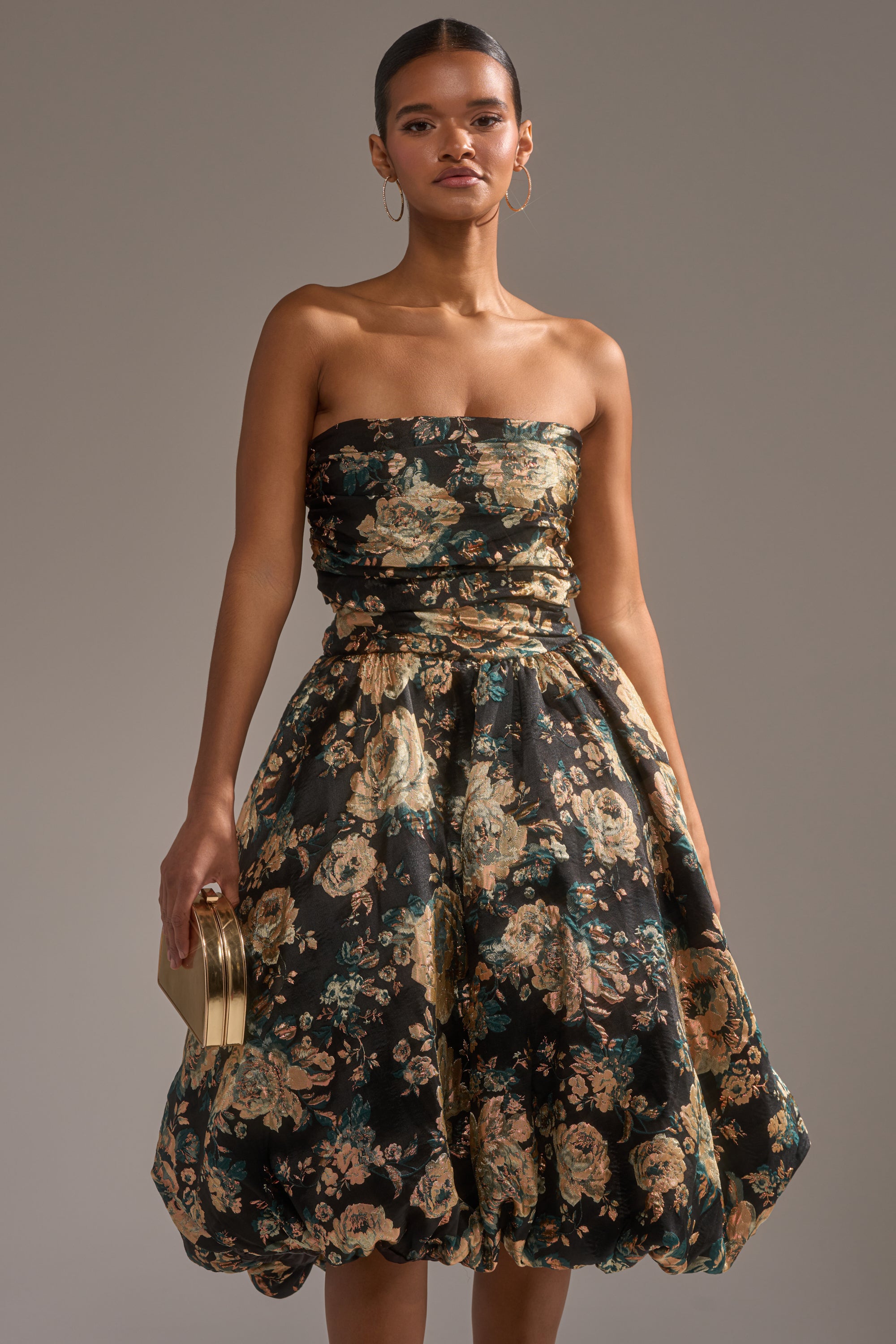 A woman wears the DRESSED TO IMPRESS BROCADE MIDI DRESS, featuring a strapless floral design and full skirt. She accessorizes with hoop earrings, a gold clutch, and has her hair pulled back, posing against a plain background. 2
