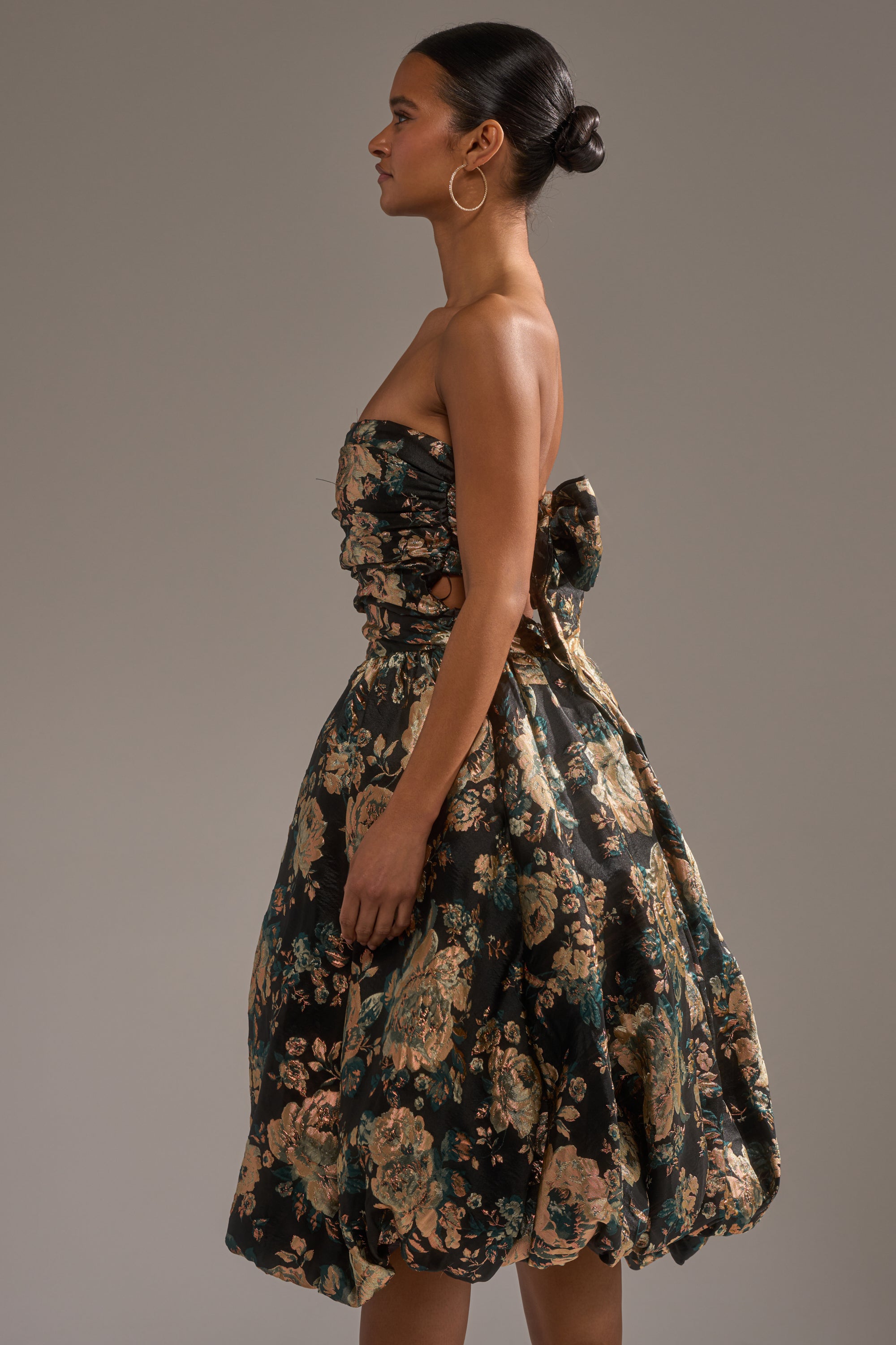 A woman in profile wears the DRESSED TO IMPRESS BROCADE MIDI DRESS, featuring a strapless top, floral brocade full skirt, and large back bow. Her hair is in a sleek bun with hoop earrings against a neutral background. 3