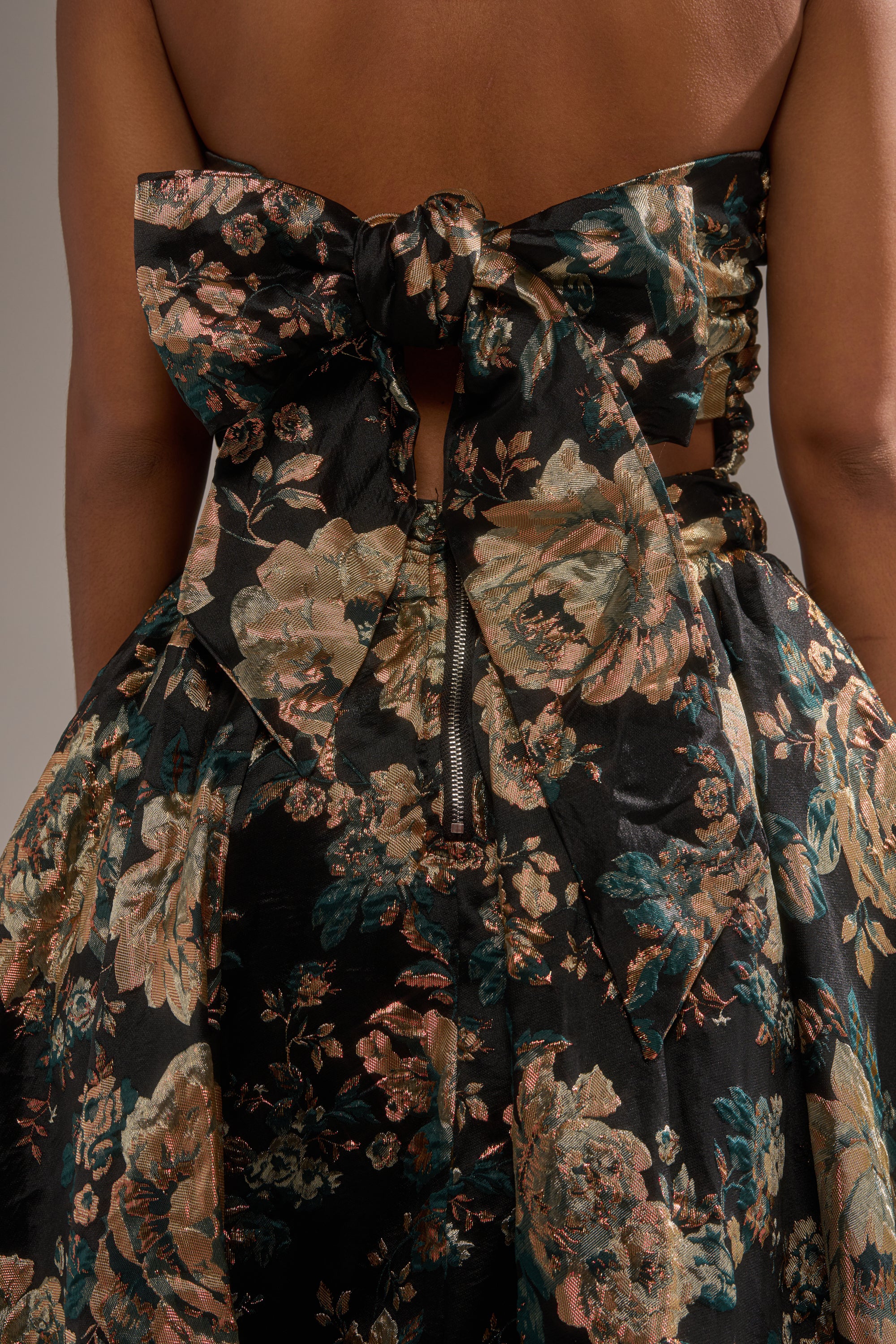 A close-up of a person wearing the DRESSED TO IMPRESS BROCADE MIDI DRESS, featuring black floral brocade with metallic gold, teal, and bronze flowers, a large back bow, and center zipper—ideal for holiday parties. 7