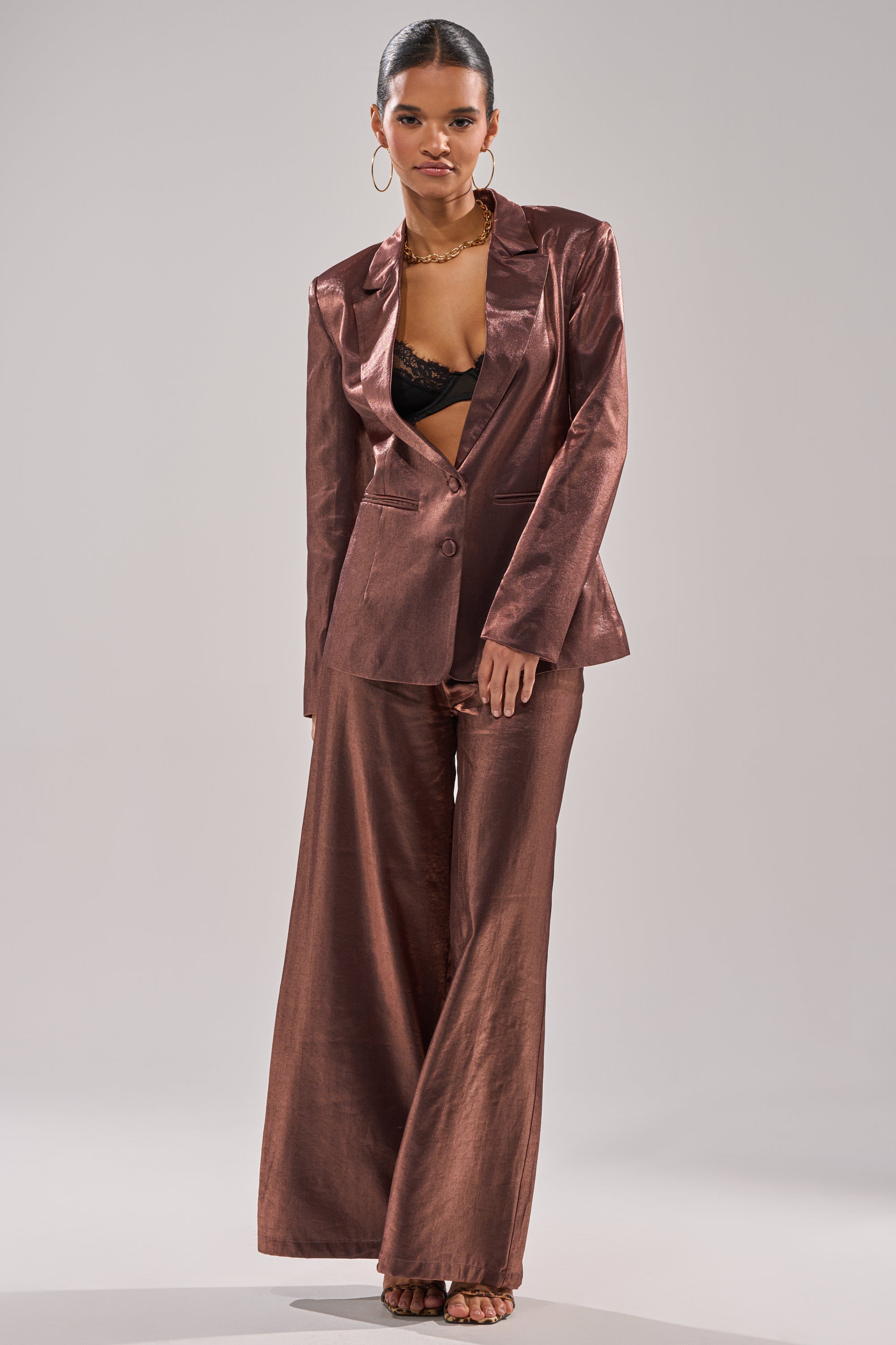 A person stands confidently in the STAY CLOSE IRIDESCENT TROUSER, a rose-brown metallic taffeta wide-leg pant, paired with an open blazer, black lace bralette, hoop earrings, gold necklace, and heeled sandals against a plain light background. 0