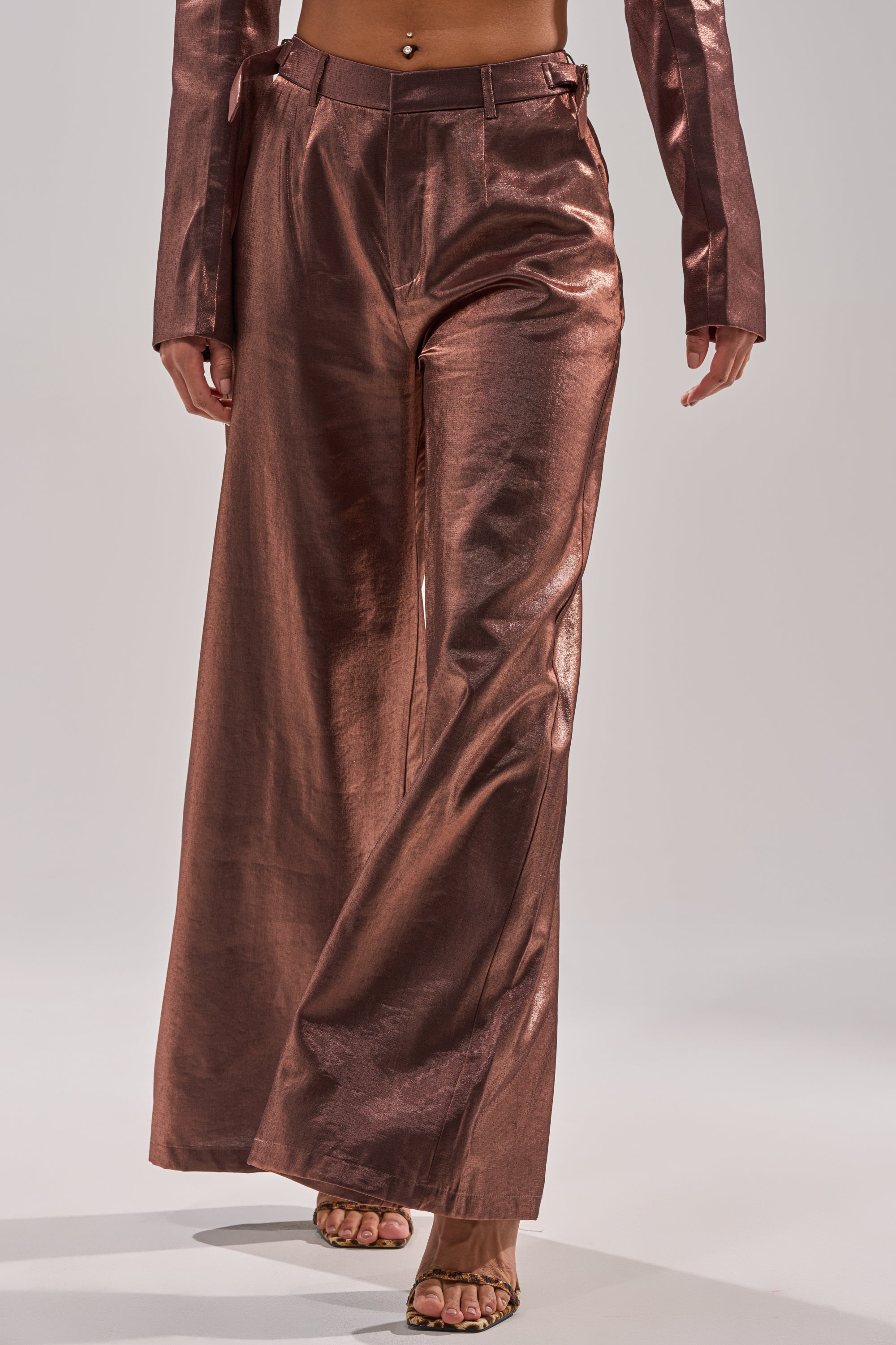A person stands against a plain background, showing only the lower torso down while wearing the STAY CLOSE IRIDESCENT TROUSER—a high-waisted, metallic taffeta, wide-leg pant—paired with open-toe sandals and a matching top. 1
