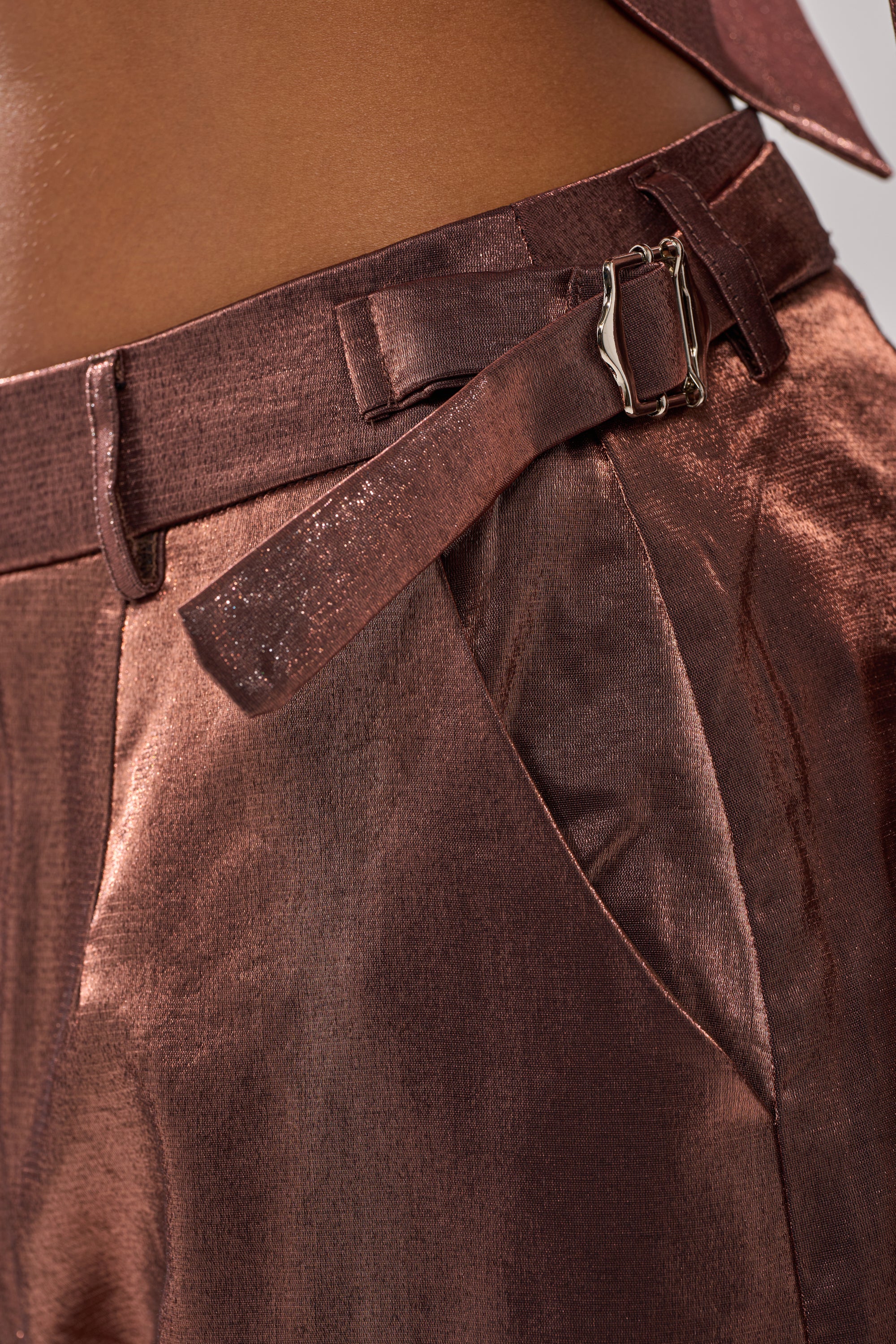 Close-up of a person wearing the STAY CLOSE IRIDESCENT TROUSER, featuring shiny metallic taffeta fabric, a wide-leg silhouette, belt loops, a side pocket, and a matching fabric belt with a silver buckle. 8