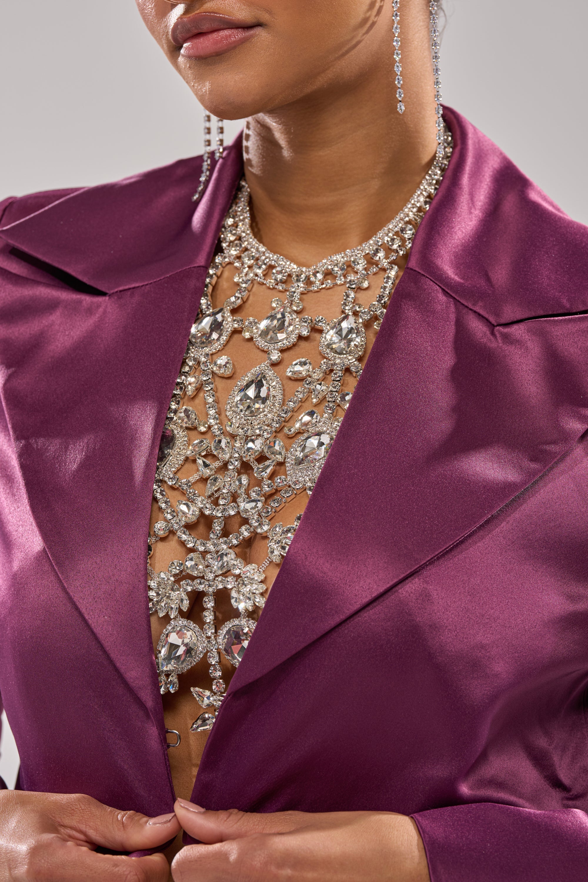 A woman wears a shiny purple blazer accented with statement jewelry—a dazzling IN YOUR DREAMS BODY CHAIN draping her chest, long dangling earrings, and sparkling rhinestone accessories. Only her lower face is visible. 1