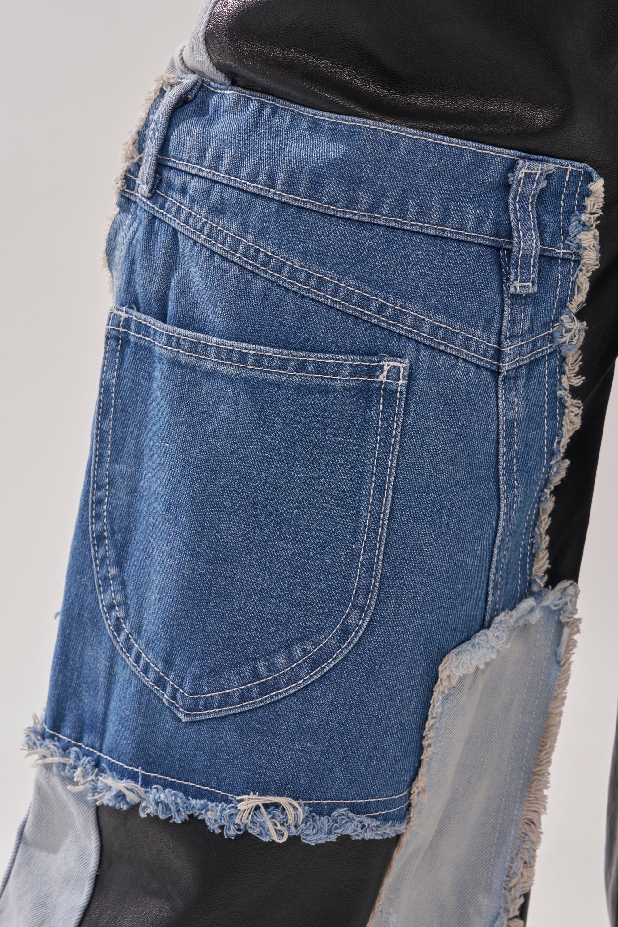 Close-up of TOO MUCH GOING ON RELAXED DENIM—denim fabric with a large pocket on black leather, frayed edges, and bold stitching, showcasing a street-style inspired patchwork design. 10