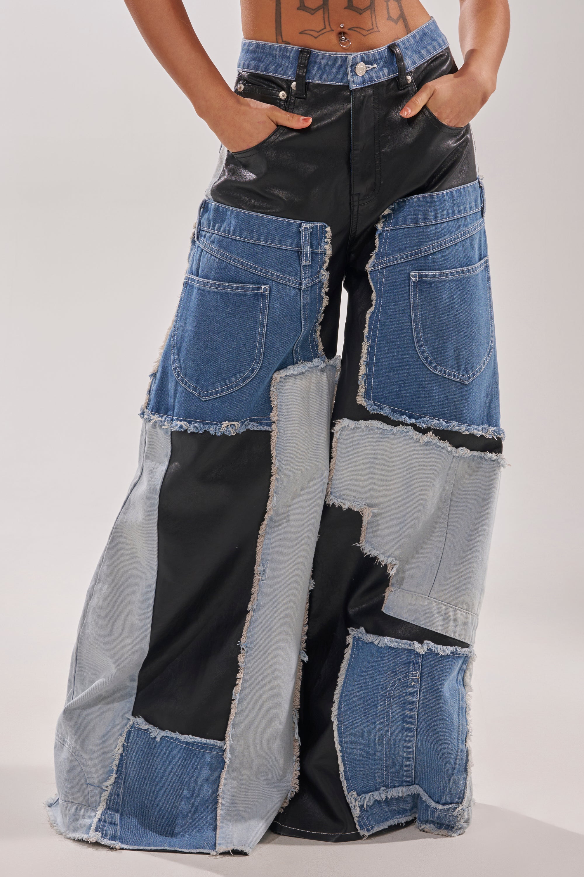 A person showcases street style in the TOO MUCH GOING ON RELAXED DENIM: wide-leg, blue and black patchwork jeans with faux leather panels, frayed seams, visible pocket details, and hands tucked into the front pockets. 2