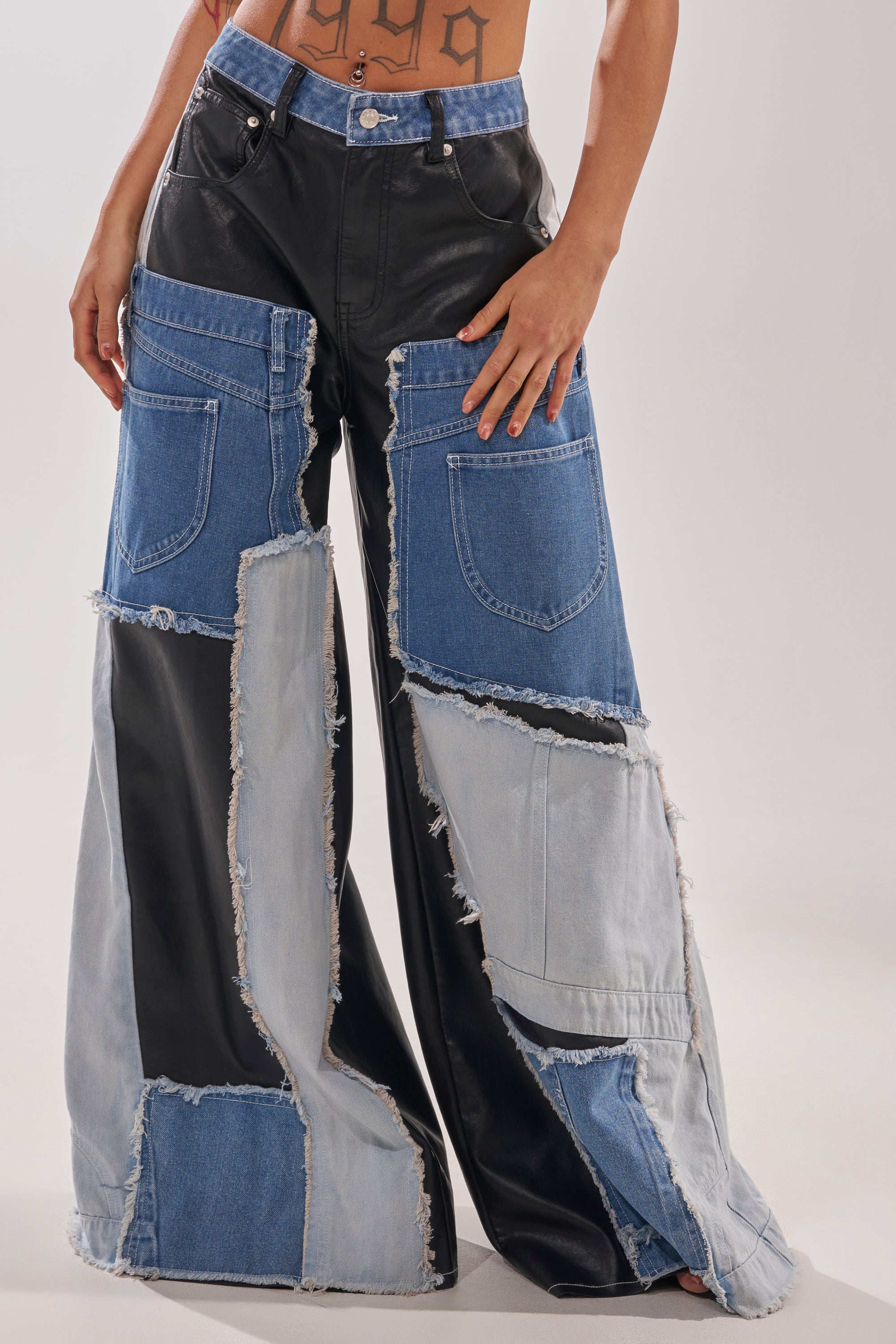 Turning heads in the TOO MUCH GOING ON RELAXED DENIM, this person channels street style with wide-leg patchwork pants featuring frayed edges, showing off midriff tattoos while resting a hand on their hip. 0